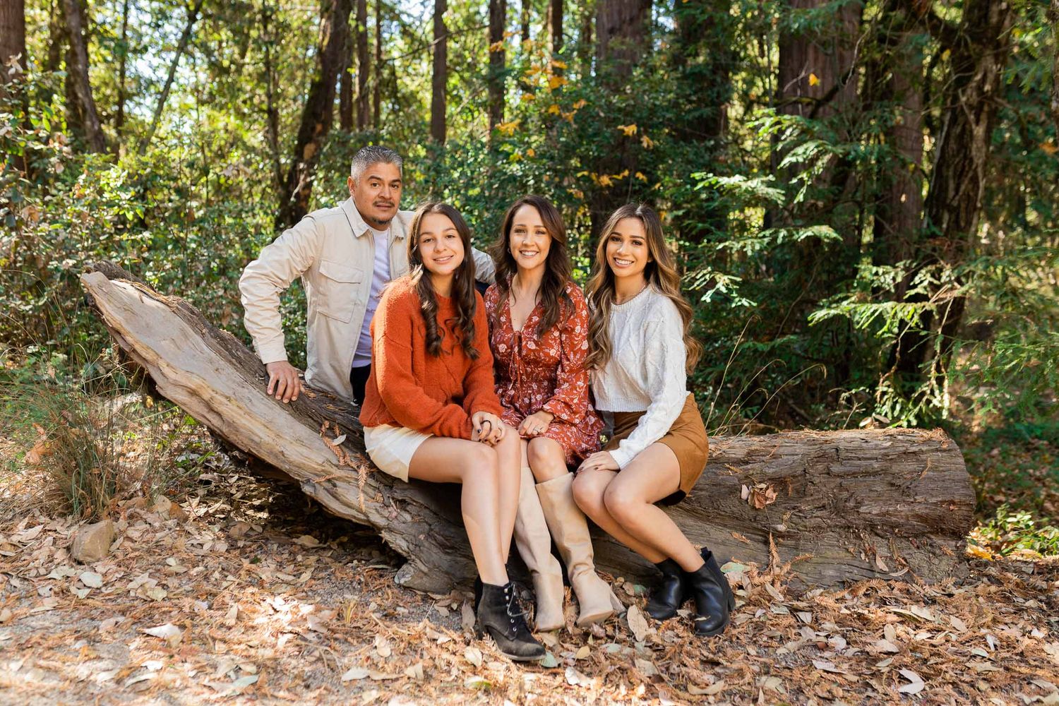 Family of four smiling together on a fallen log in a lush forest during autumn.