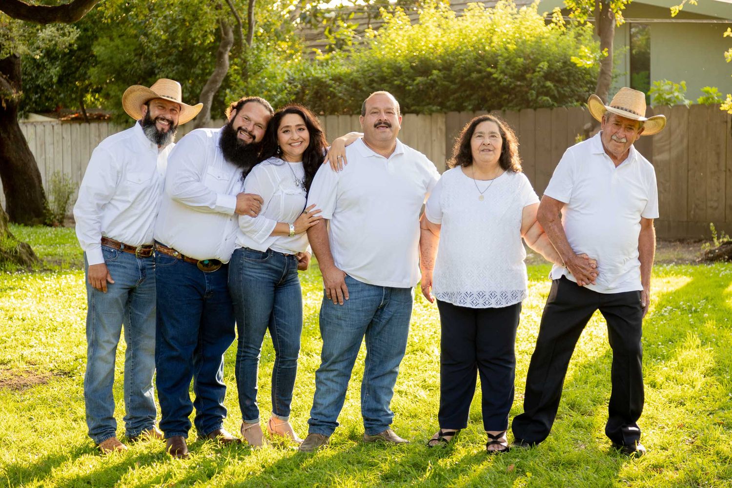 Six people in white shirts and jeans pose together smiling outdoors on green grass with trees and a wooden fence.