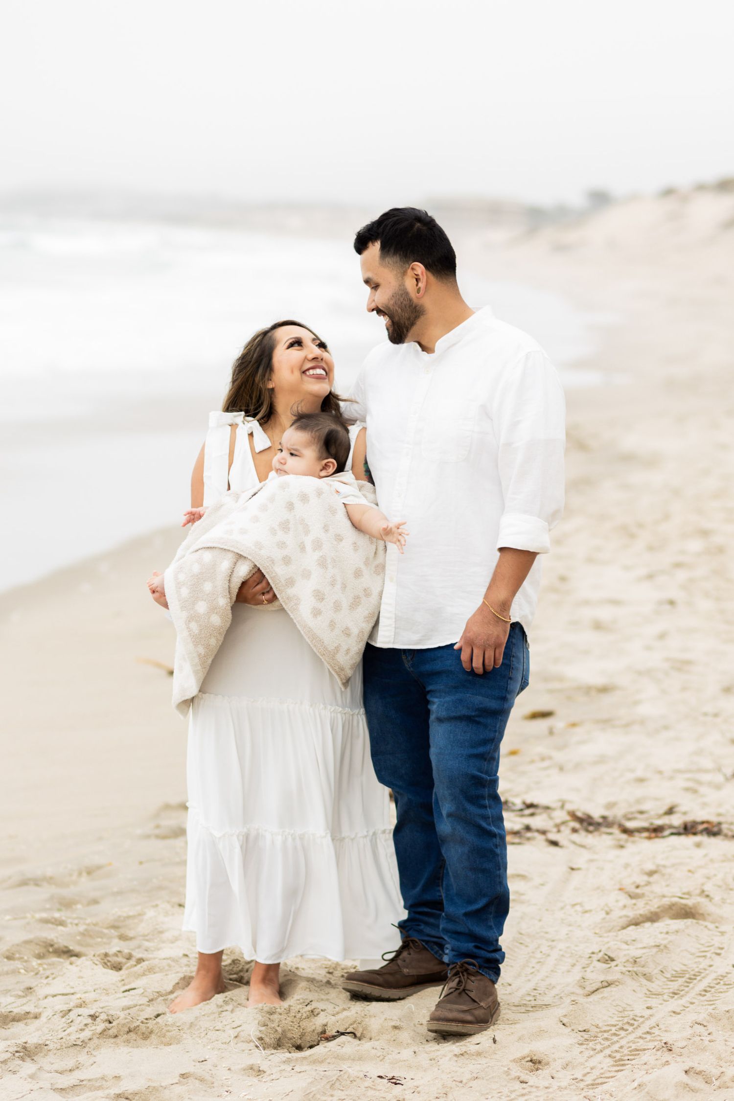 Happy couple with baby on a sandy beach, woman in white dress holding infant.
