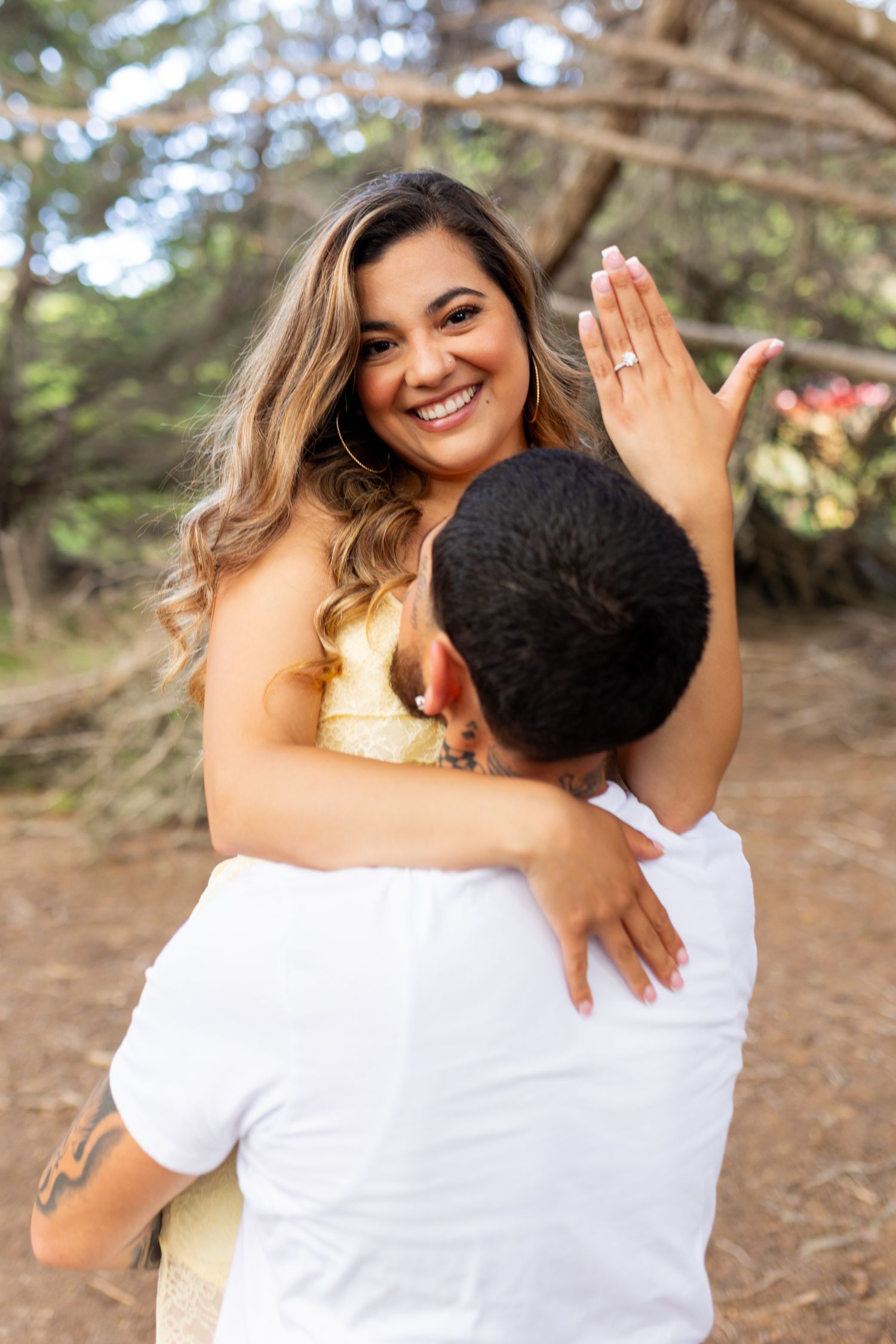A joyful woman shows off her engagement ring while being embraced by her partner outdoors among trees.