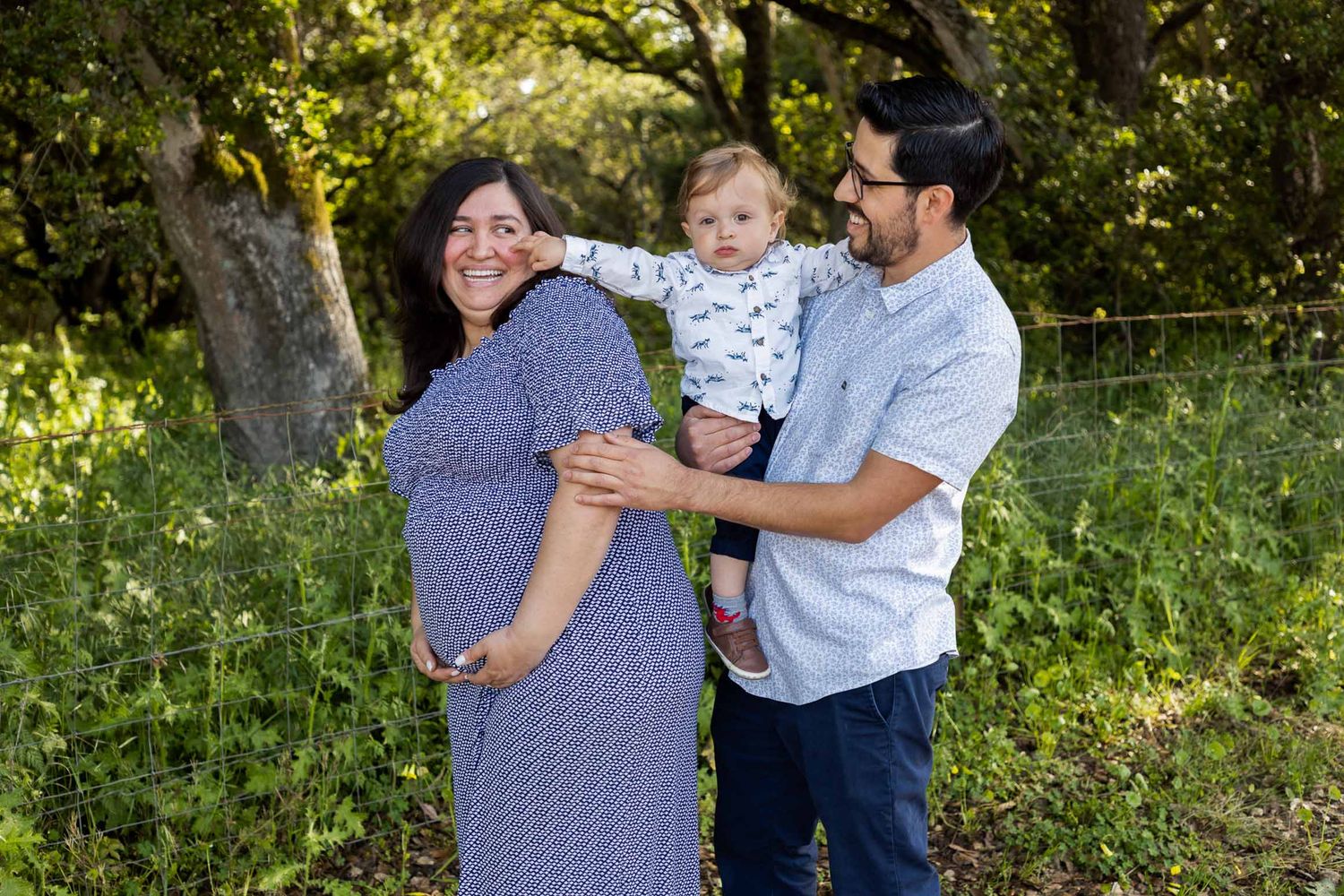 Pregnant woman in blue dress with husband holding toddler smiling outdoors in lush green nature setting.