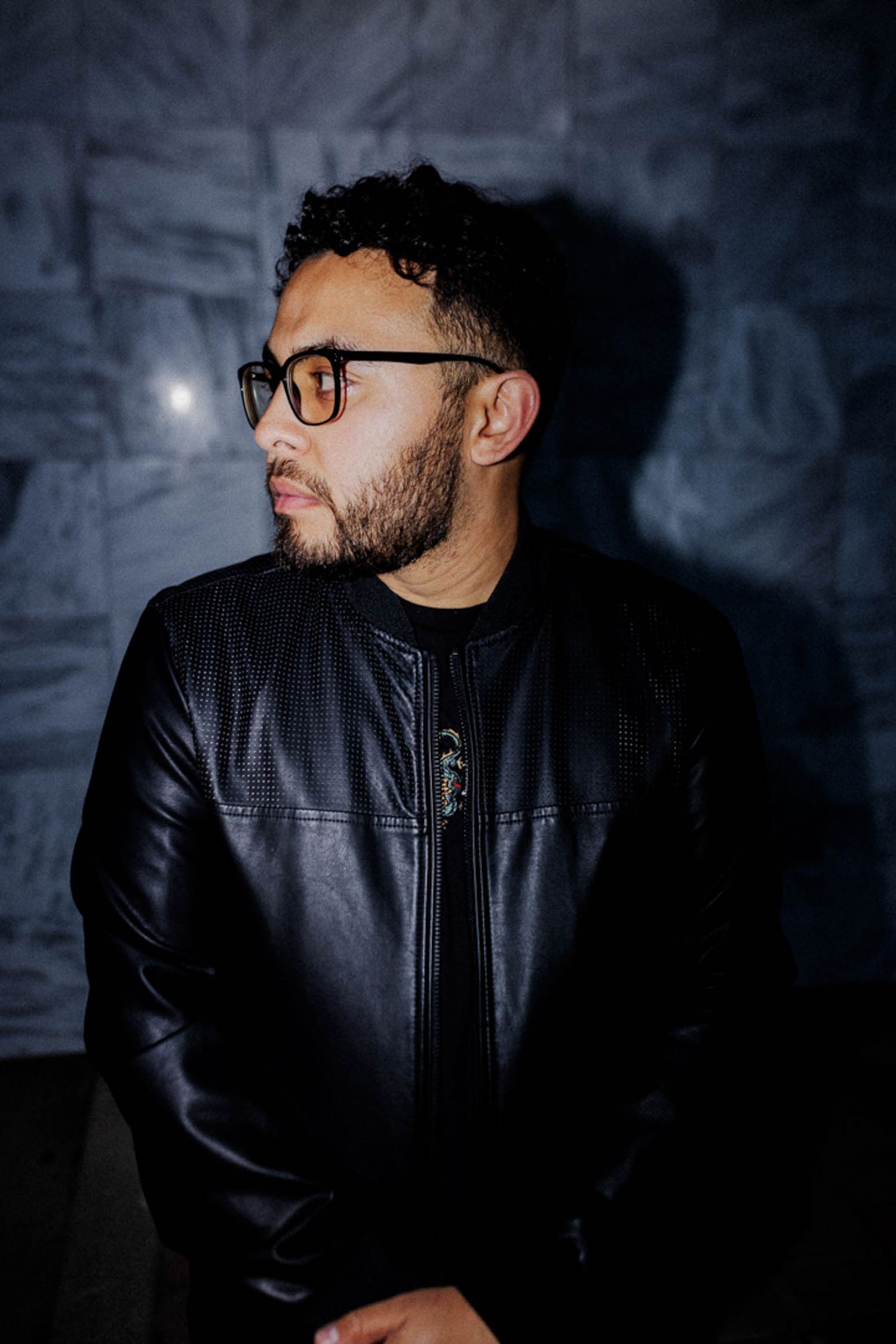 Person wearing glasses and black leather jacket poses against dark textured wall in moody lighting.