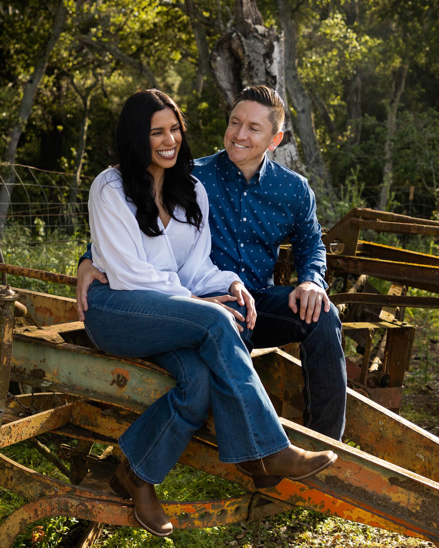 A smiling couple sits together on rustic farm equipment surrounded by lush green trees in an outdoor engagement photo.