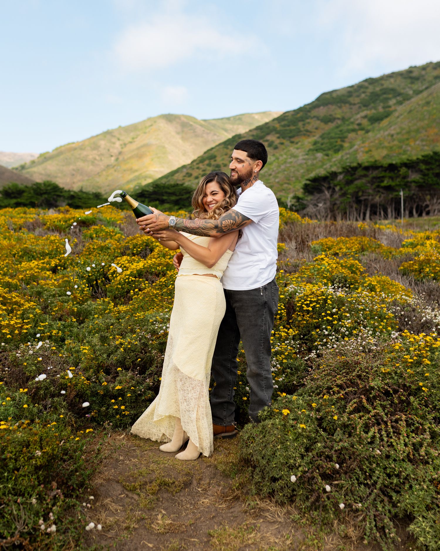 Couple celebrating in yellow wildflowers with champagne, green hills backdrop, woman in cream dress, man with tattoos.