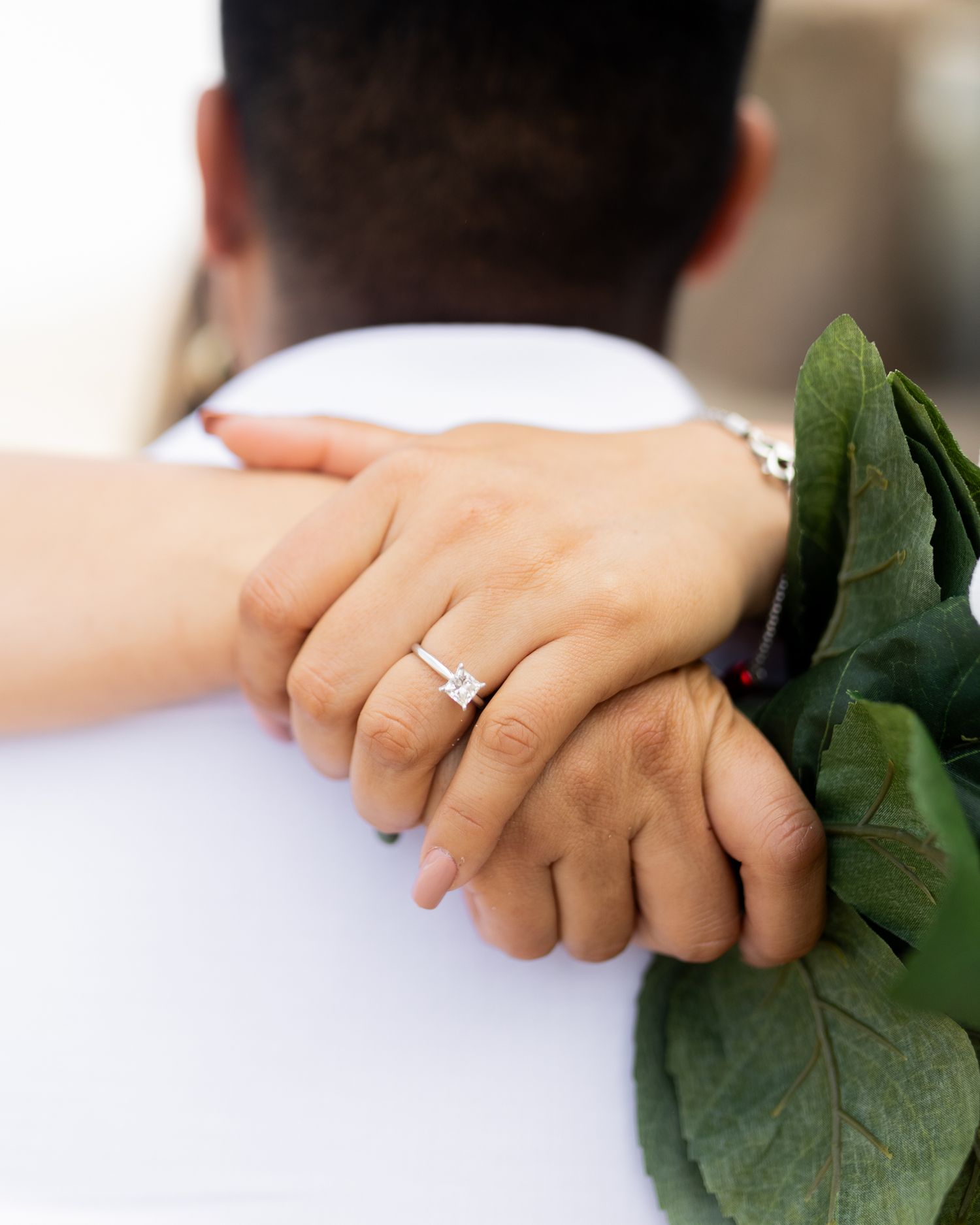 A close-up of a woman's hand with a diamond engagement ring resting on a man's shoulder while holding green leaves.