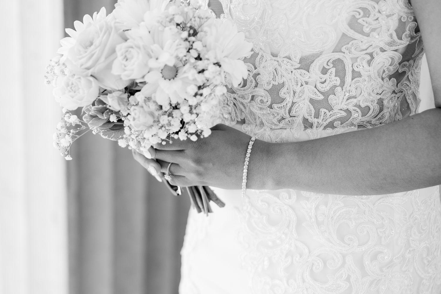 Black and white photo of a bride in lace gown holding a bouquet of daisies and roses with a pearl bracelet.