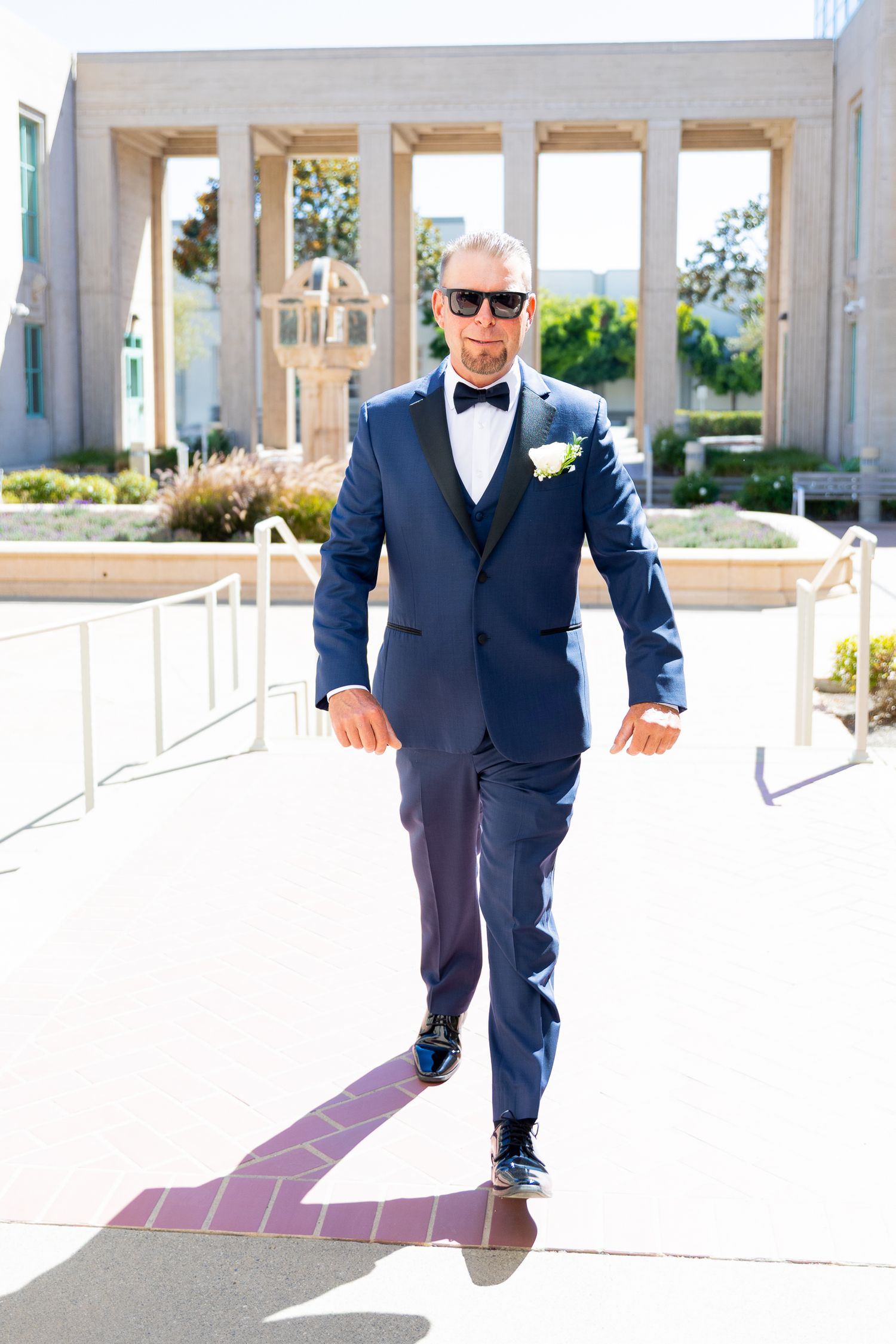 Groom in navy blue tuxedo with bow tie and boutonniere walks confidently outdoors on sunny wedding day.