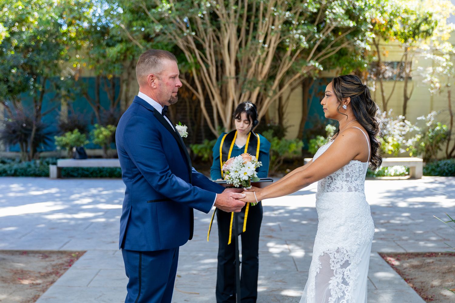 Couple exchanges vows outdoors holding hands, officiant in yellow stole behind them, lush green trees in background.