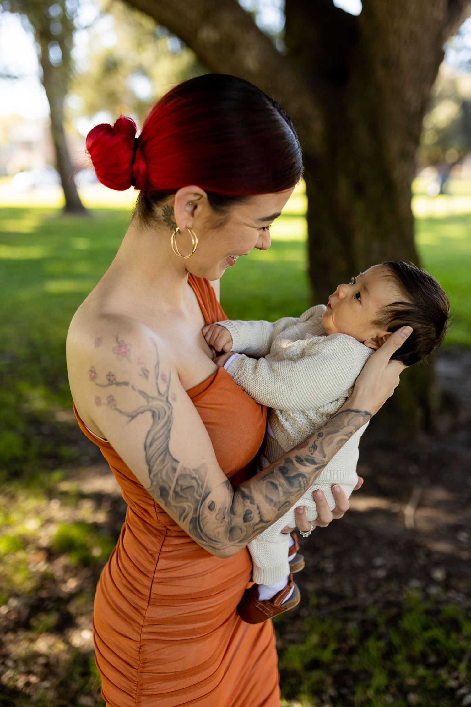 Tattooed woman with red hair in orange dress lovingly holds newborn baby outdoors in sunny green park.