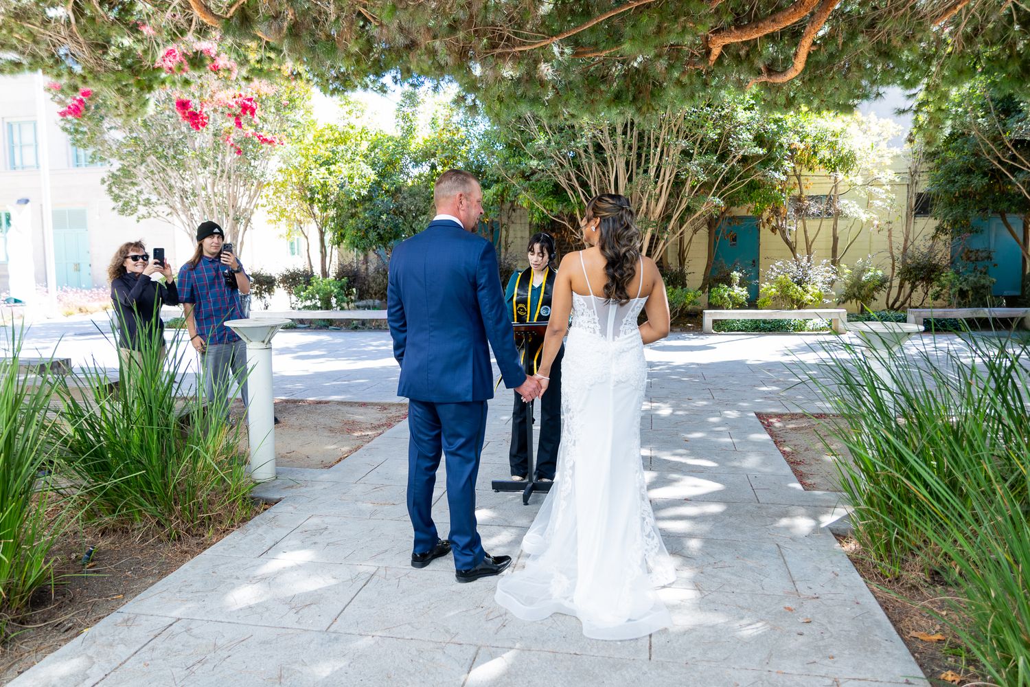 Bride and groom hold hands outdoors at a garden wedding ceremony while a photographer captures the moment.