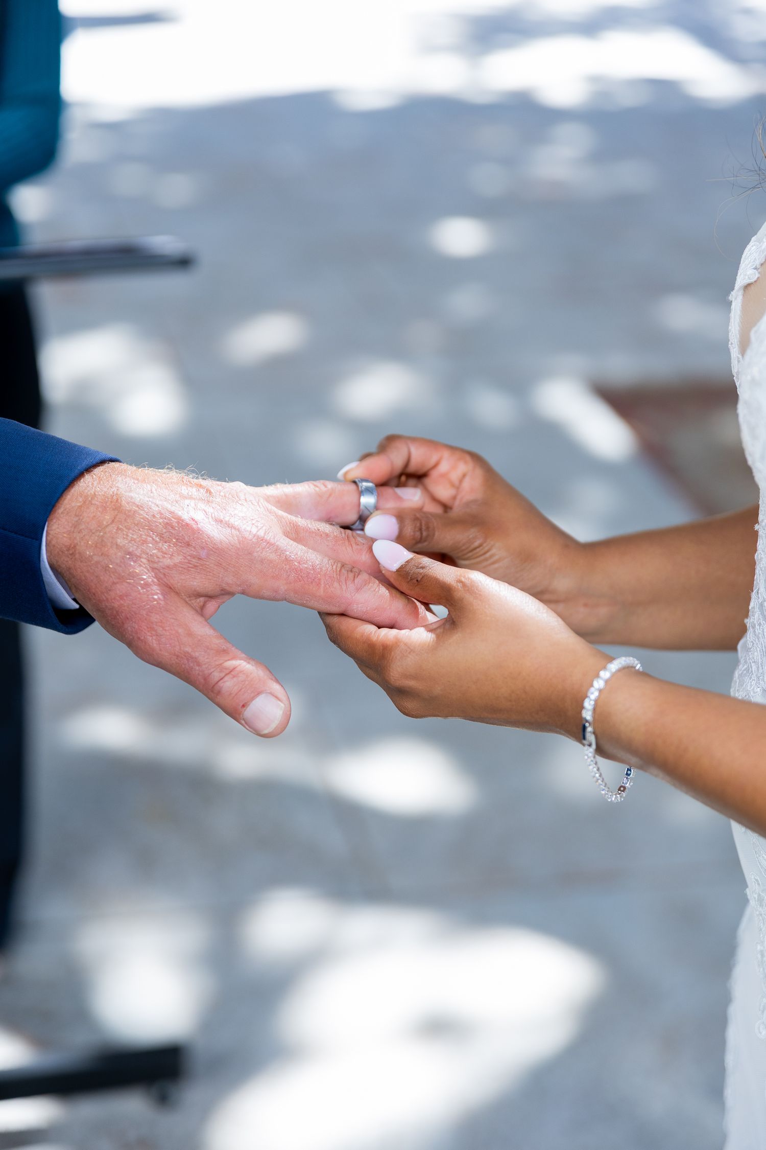 Bride placing a wedding ring on groom's finger during outdoor ceremony, close-up of hands with bracelet.
