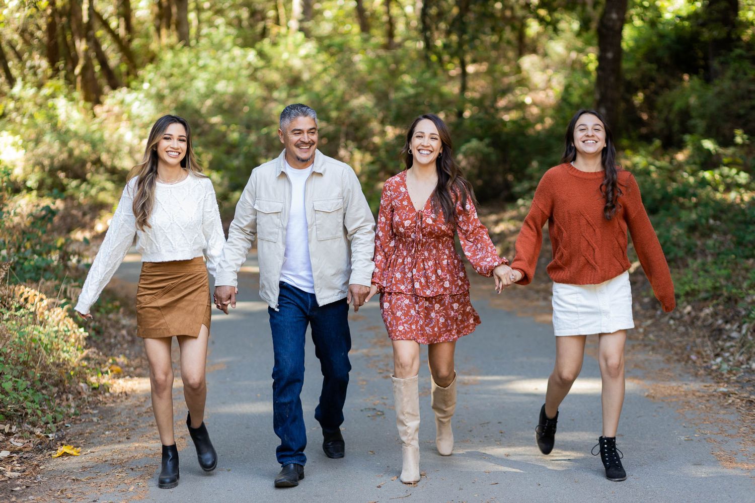Family of four holding hands walking down a tree-lined path in autumn outfits.