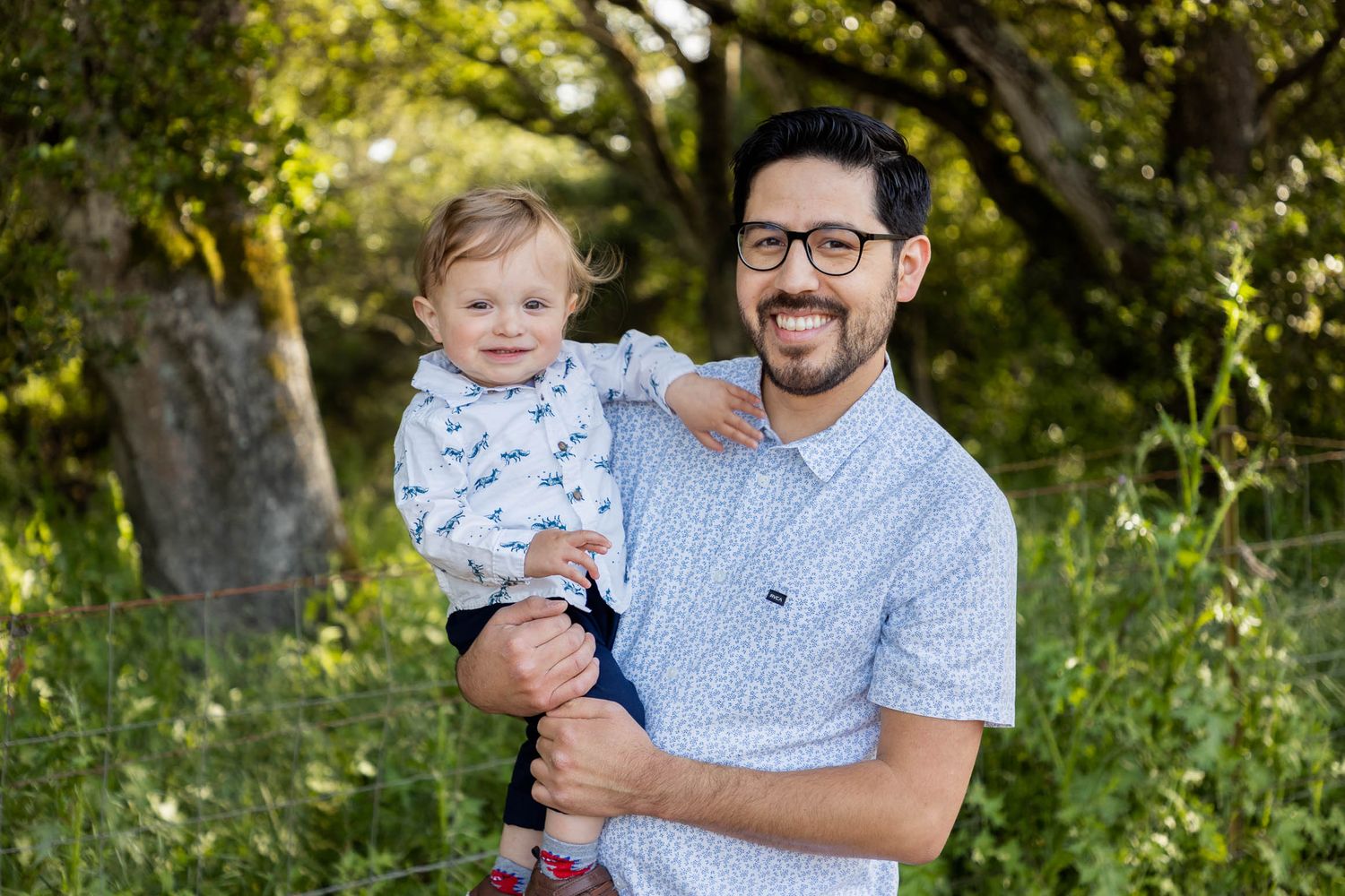 A smiling man with glasses holds a happy toddler outdoors among lush green trees in a bright family portrait.