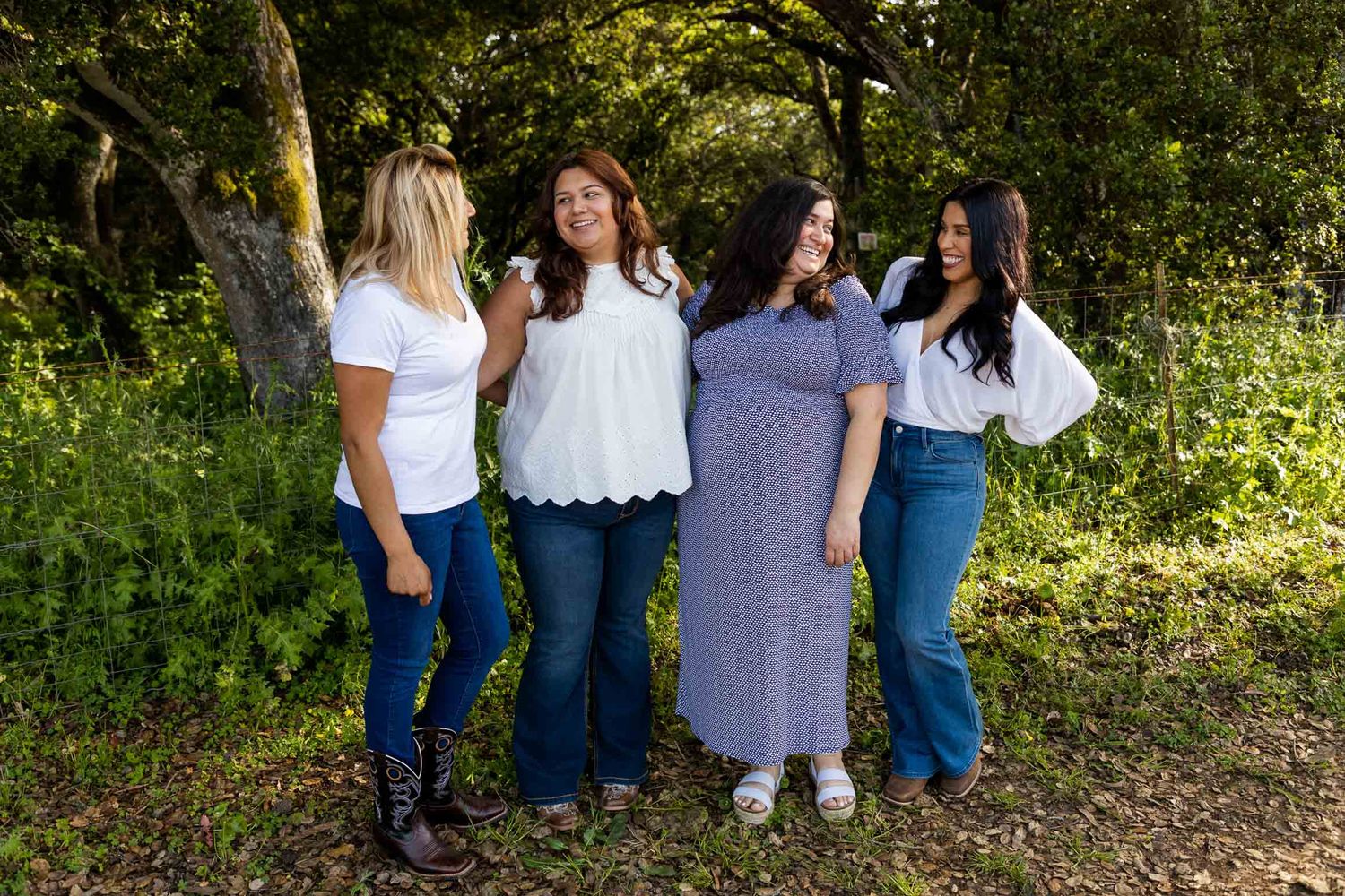 Four women smiling and posing together outdoors in a lush green wooded setting, wearing casual jeans and tops.