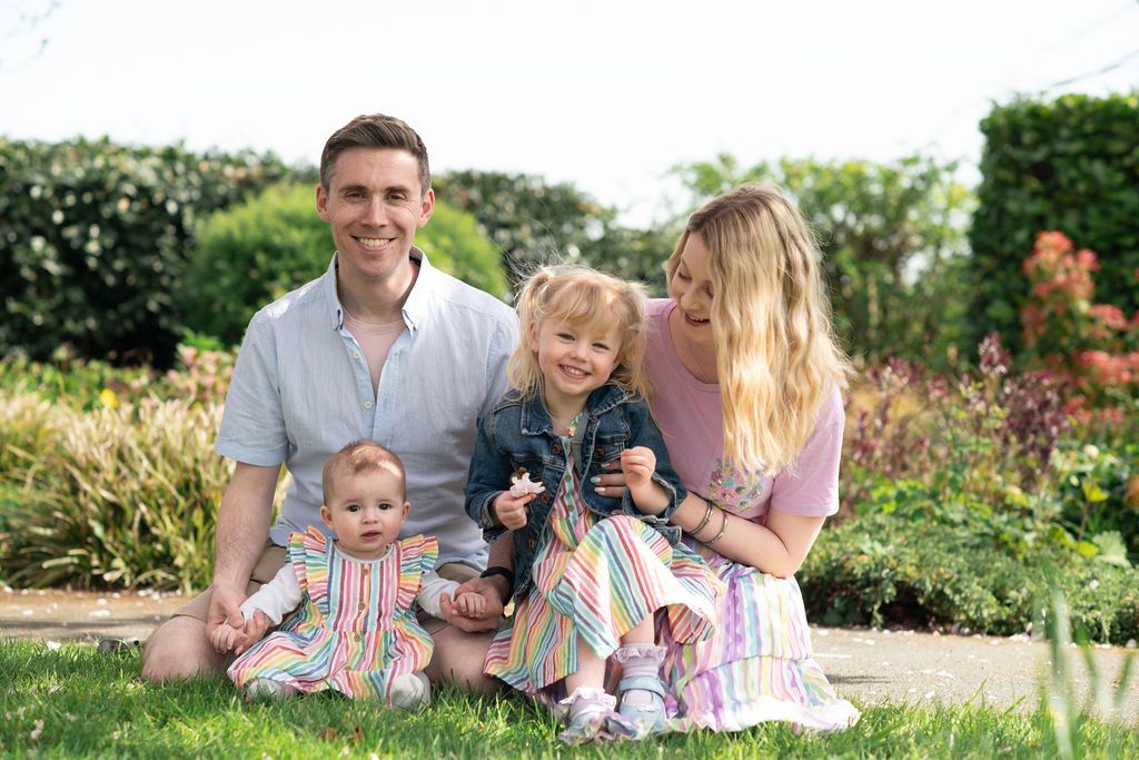 Happy family of four posing outdoors in a lush garden, parents smiling with two young children on a sunny day.