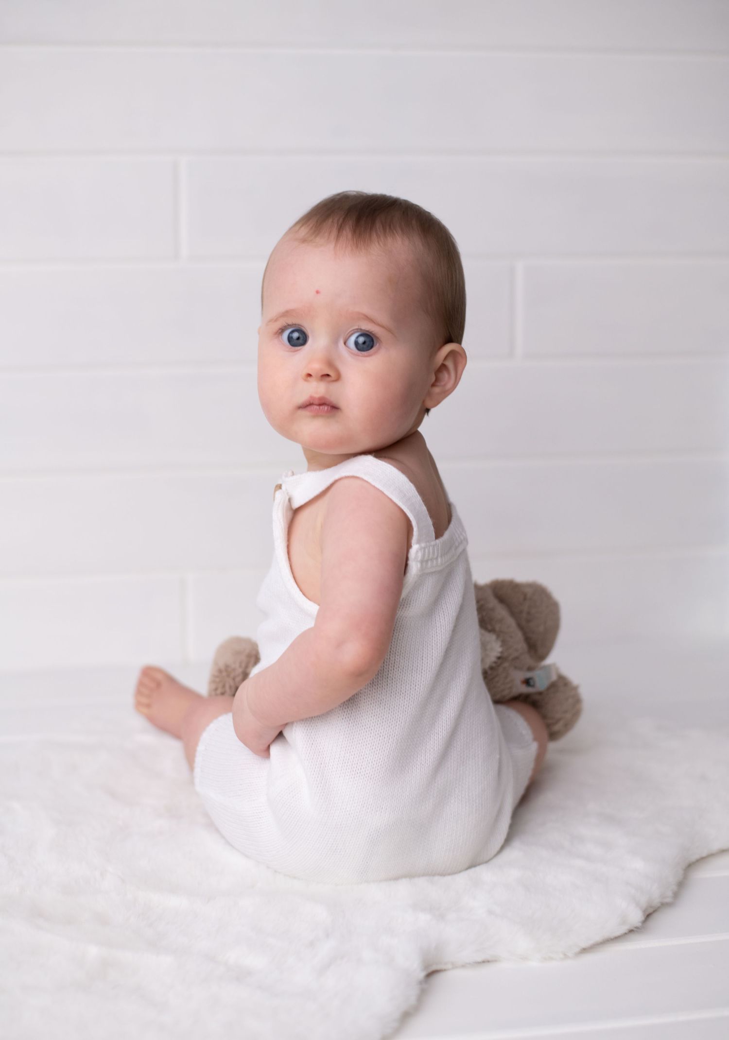 Baby wearing white onesie sits on white blanket.