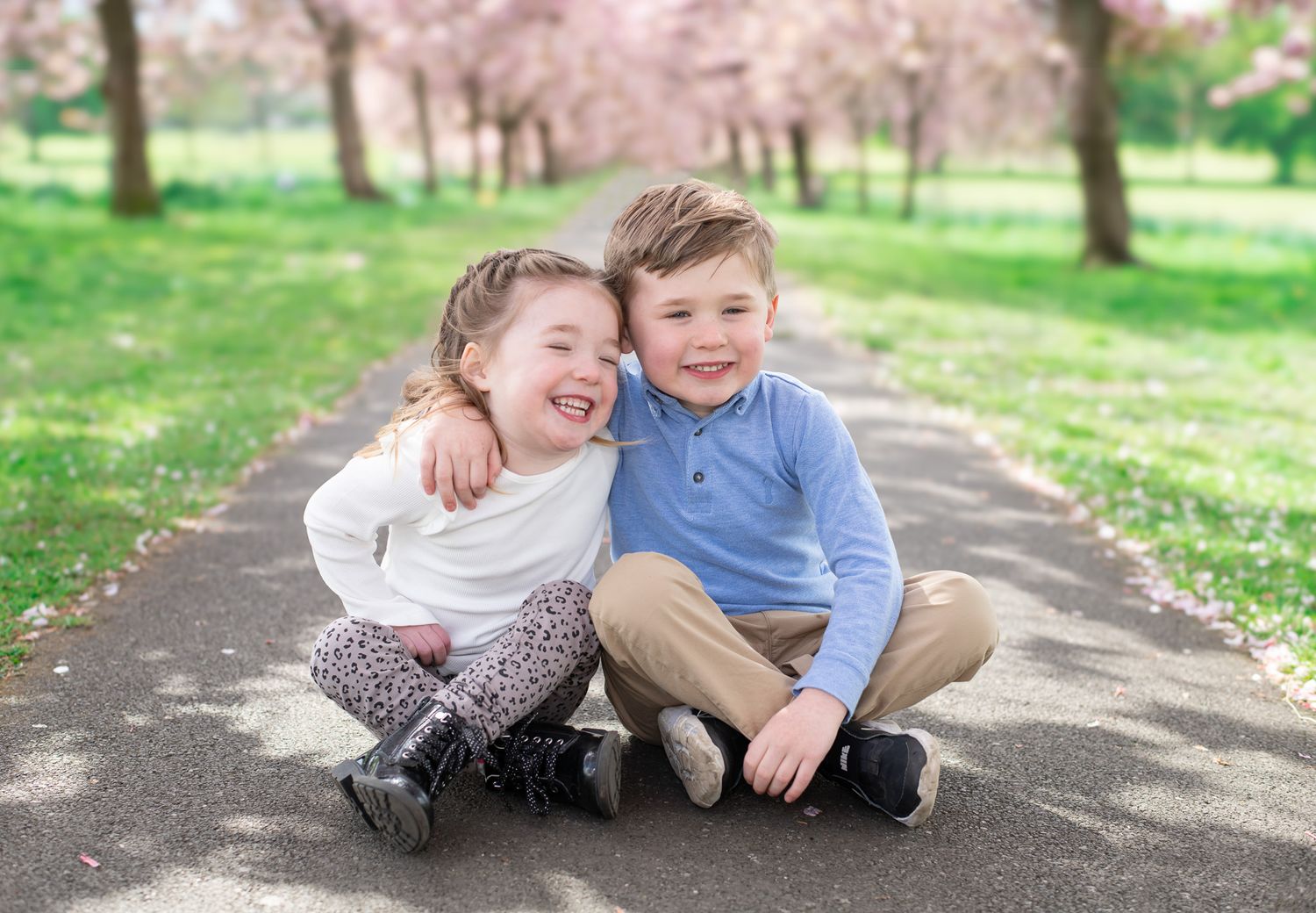 Two children sitting together and laughing on a path with blooming cherry trees in the background.
