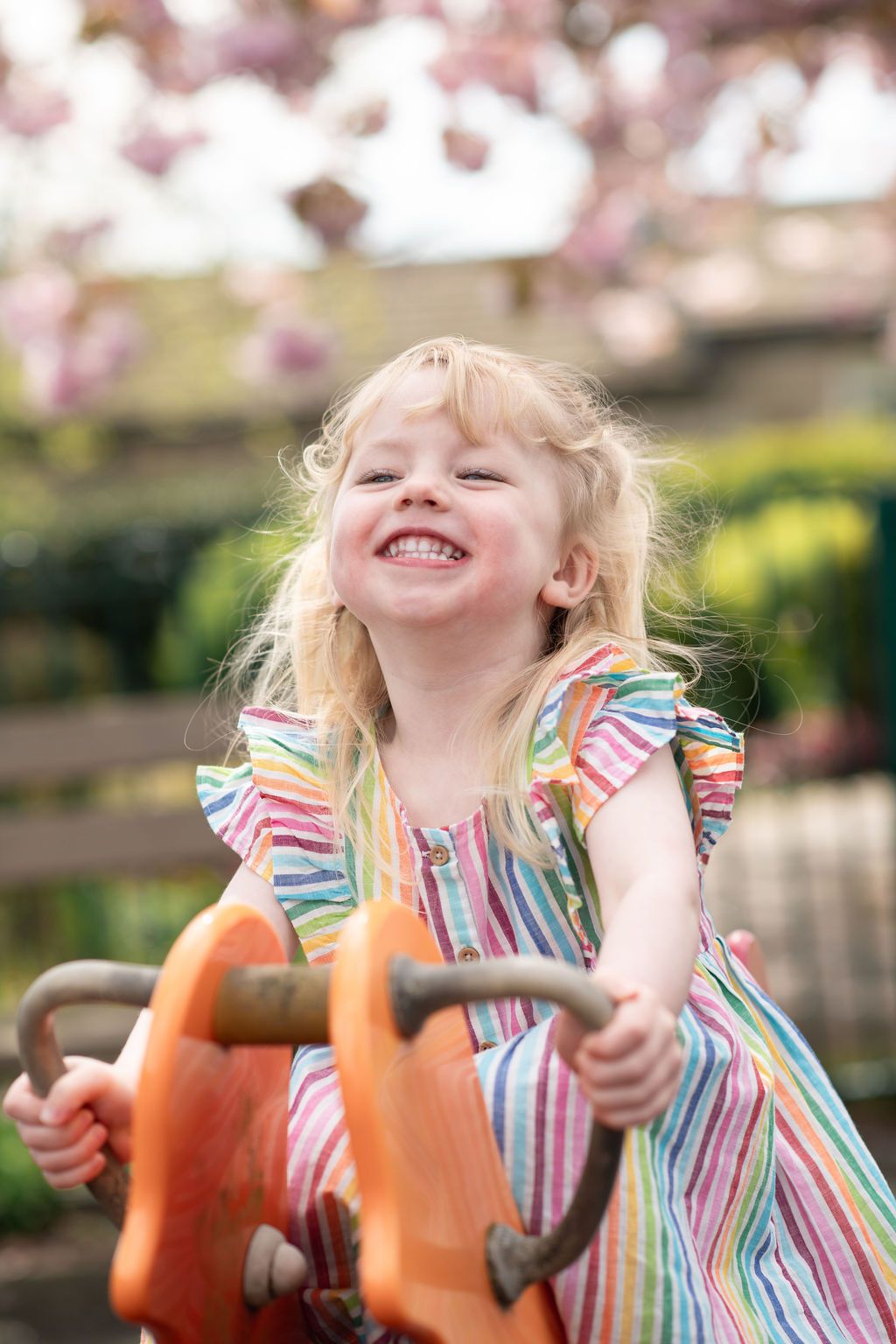 Joyful blonde toddler girl in colorful striped dress laughing on playground ride with pink cherry blossoms.