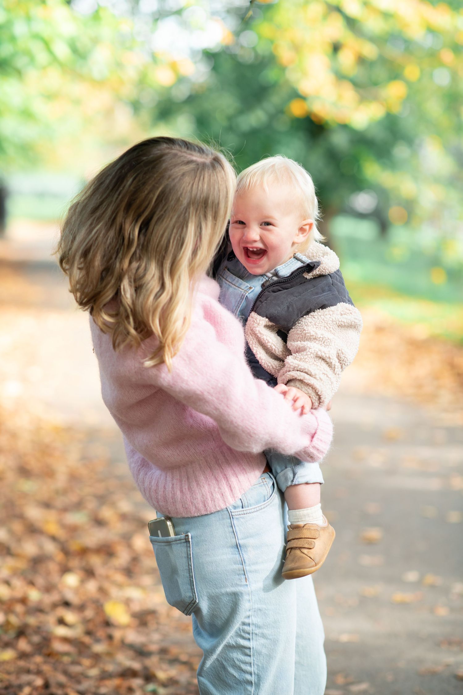 A parent in jeans and pink sweater holds a small baby in pink outfit among autumn leaves on a path.