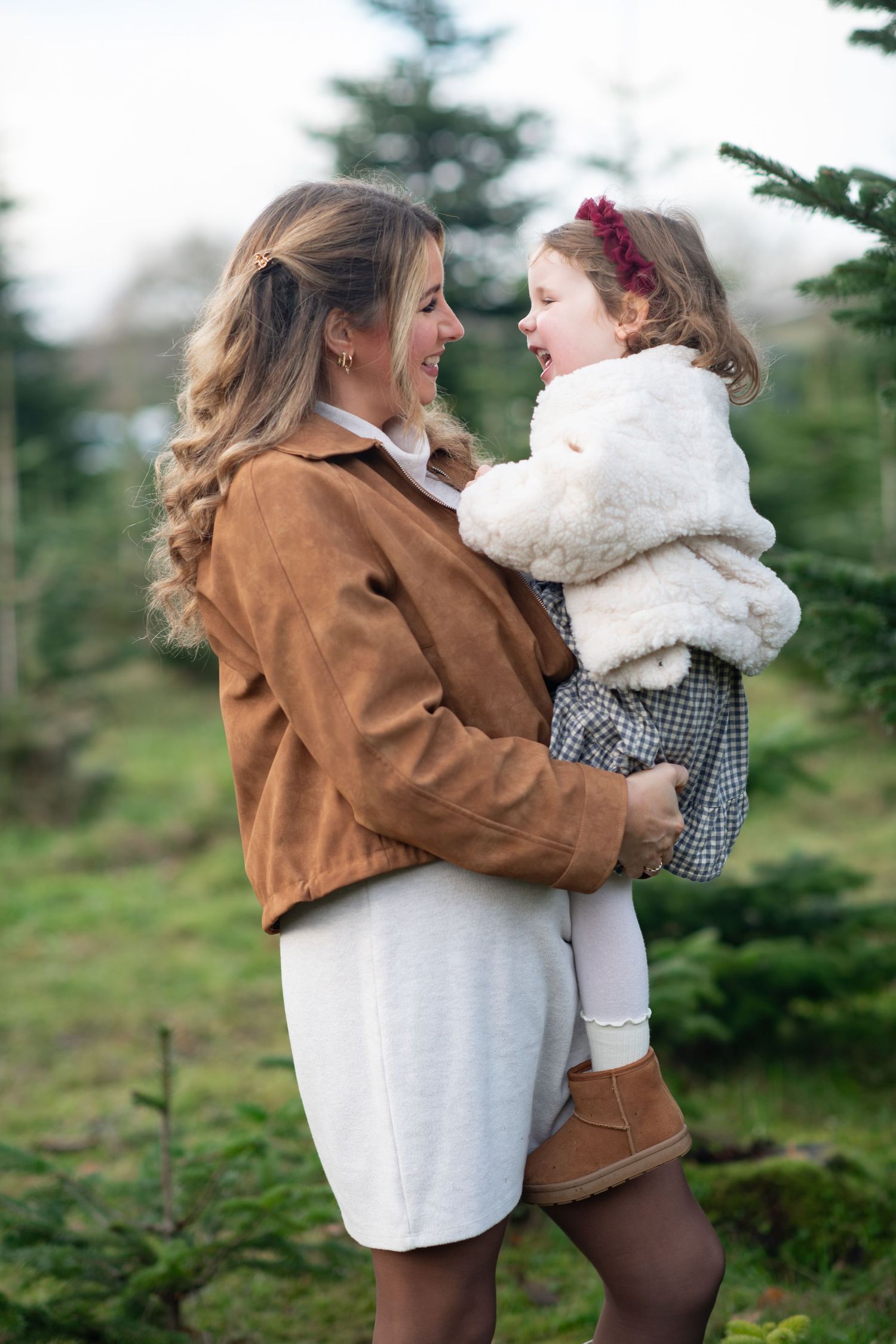 Mother and daughter share tender moments among evergreen trees in matching brown and white winter outfits.