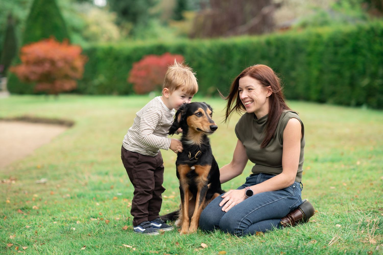 A mother and son sit with their dog in a lovely green space.
