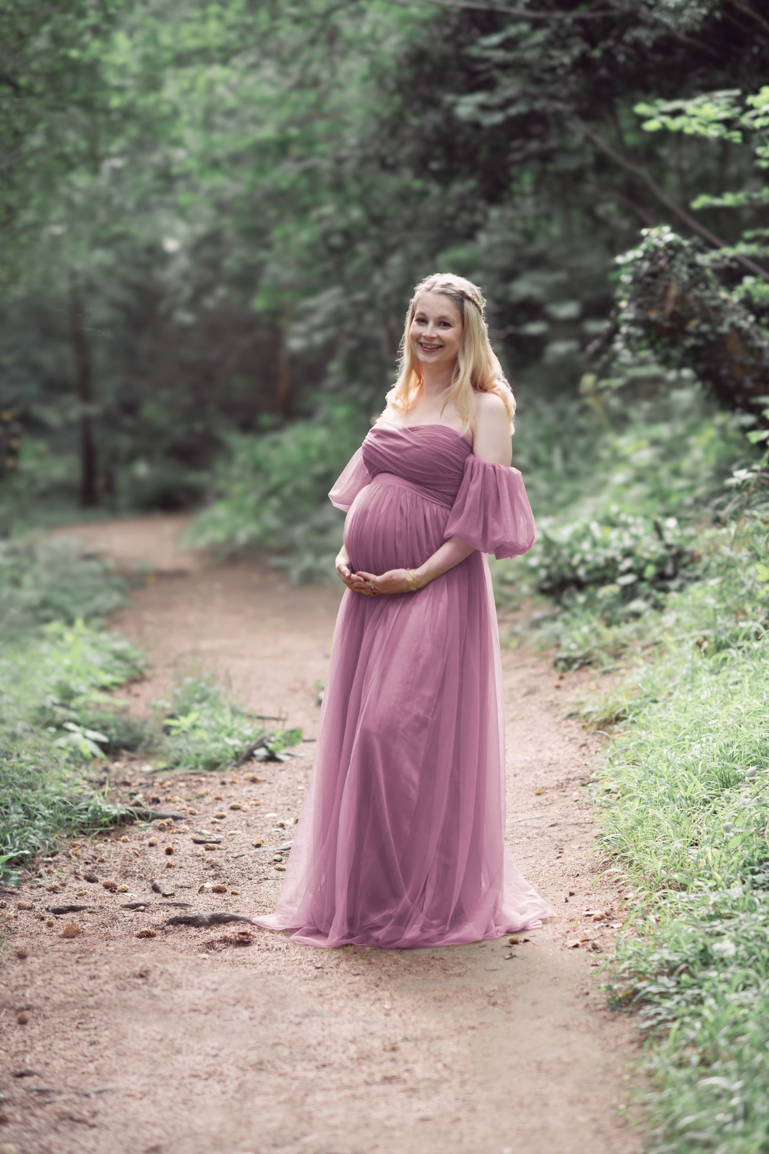 A maternity photo session on a wooded trail features a flowing mauve dress against lush green foliage.
