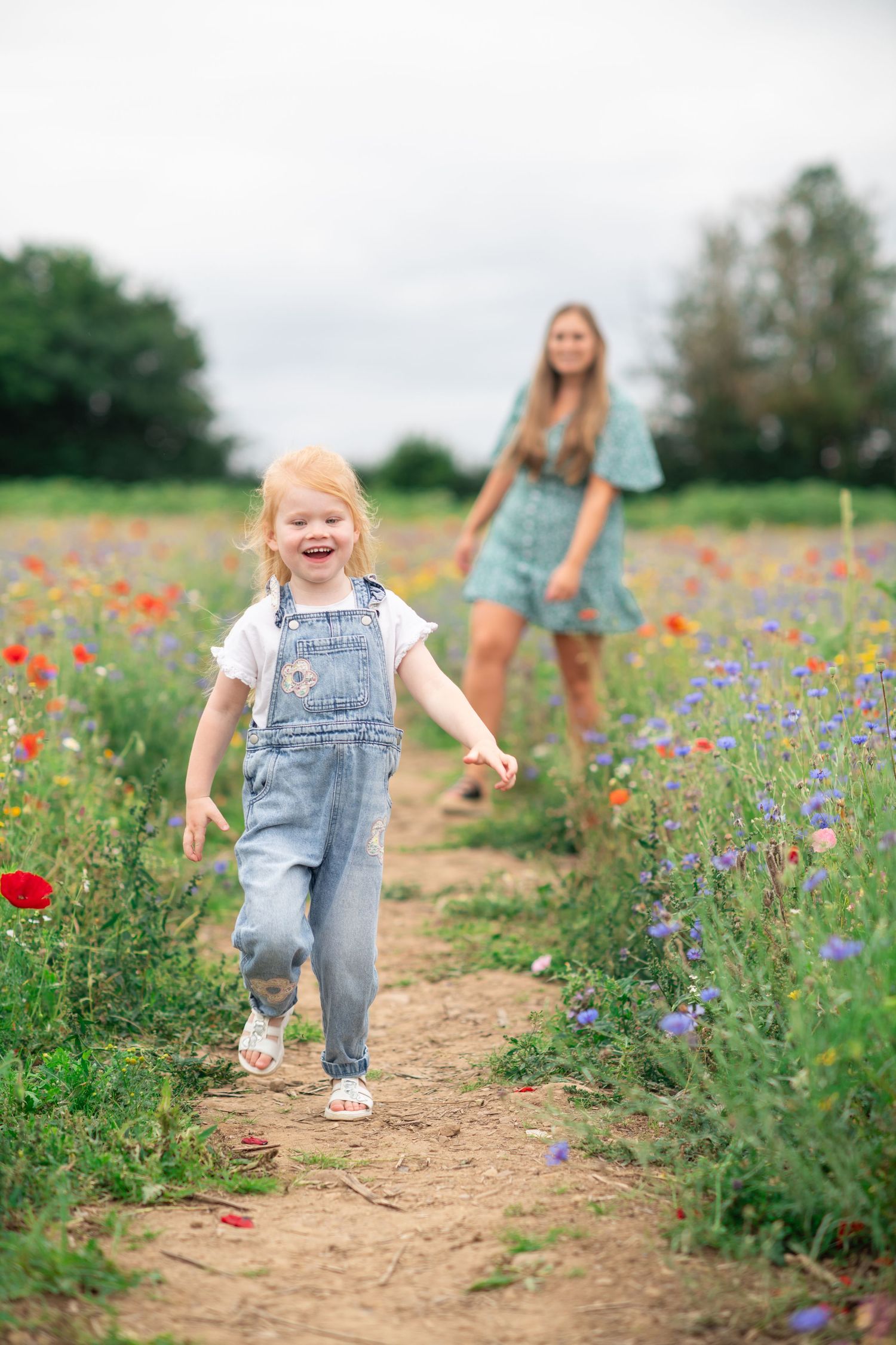 Mother and young daughter walking along a dirt path through a vibrant wildflower meadow with red poppies and purple blooms.
