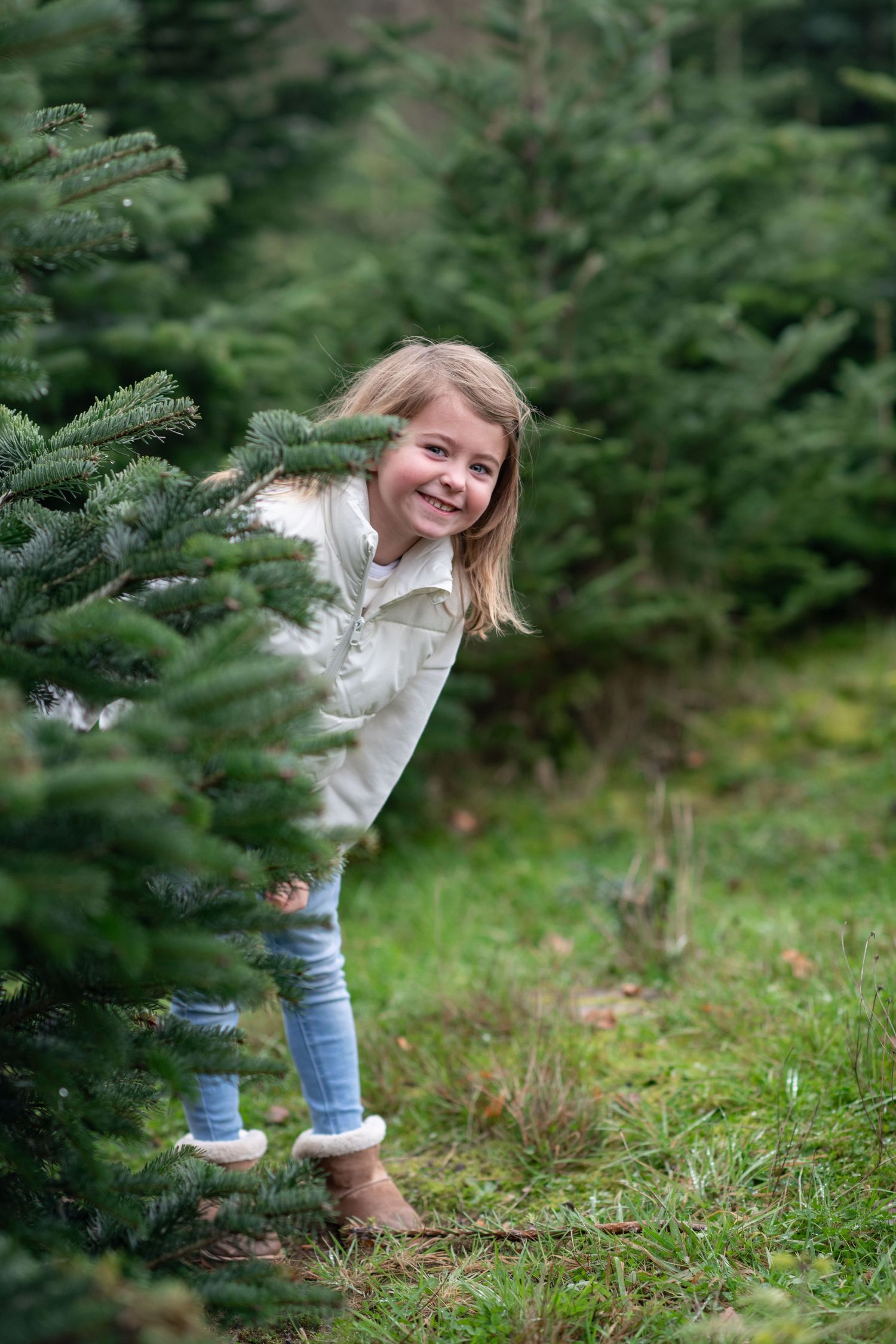 A child in jeans and a white top peeks playfully from behind a Christmas tree in an outdoor setting.