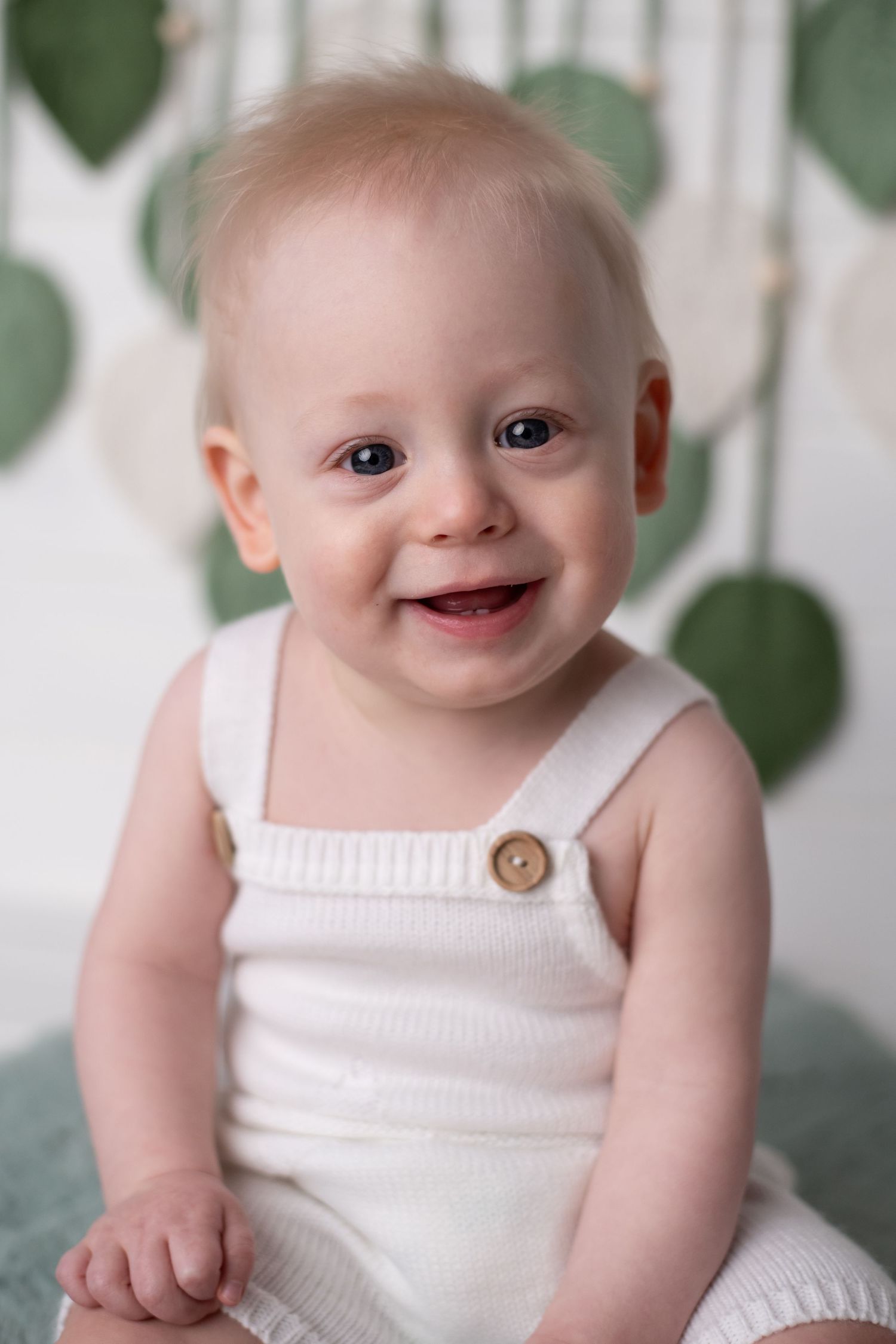 Baby wearing a white romper suit smiles brightly against green leaf backdrop.