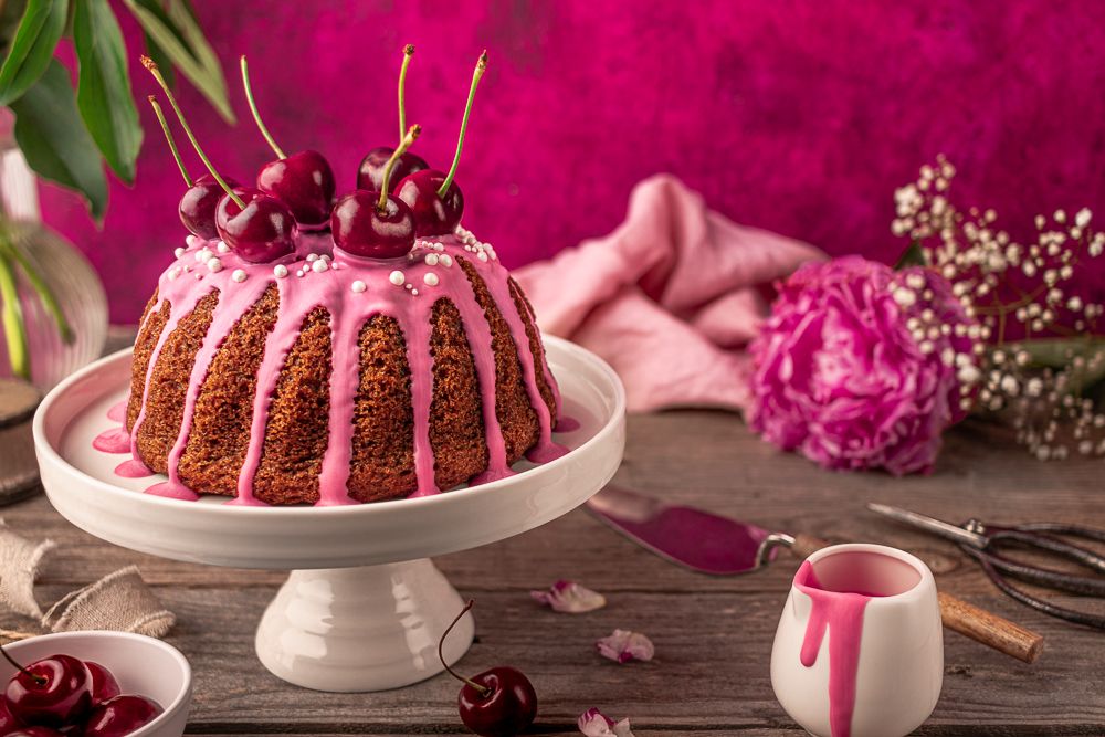 Cherry-topped bundt cake with pink glaze, flowers and a magenta backdrop, commercial food photography. Photo © Reka Peurala Three Pod Studio