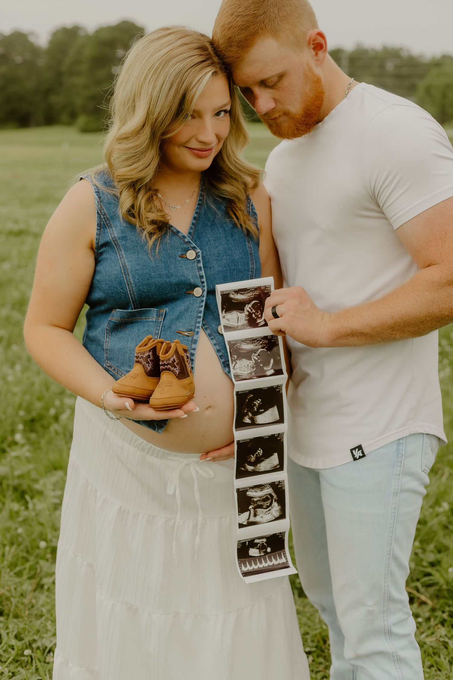 Pregnant couple outdoors holding ultrasound photos and tiny baby shoes, maternity announcement in green field.