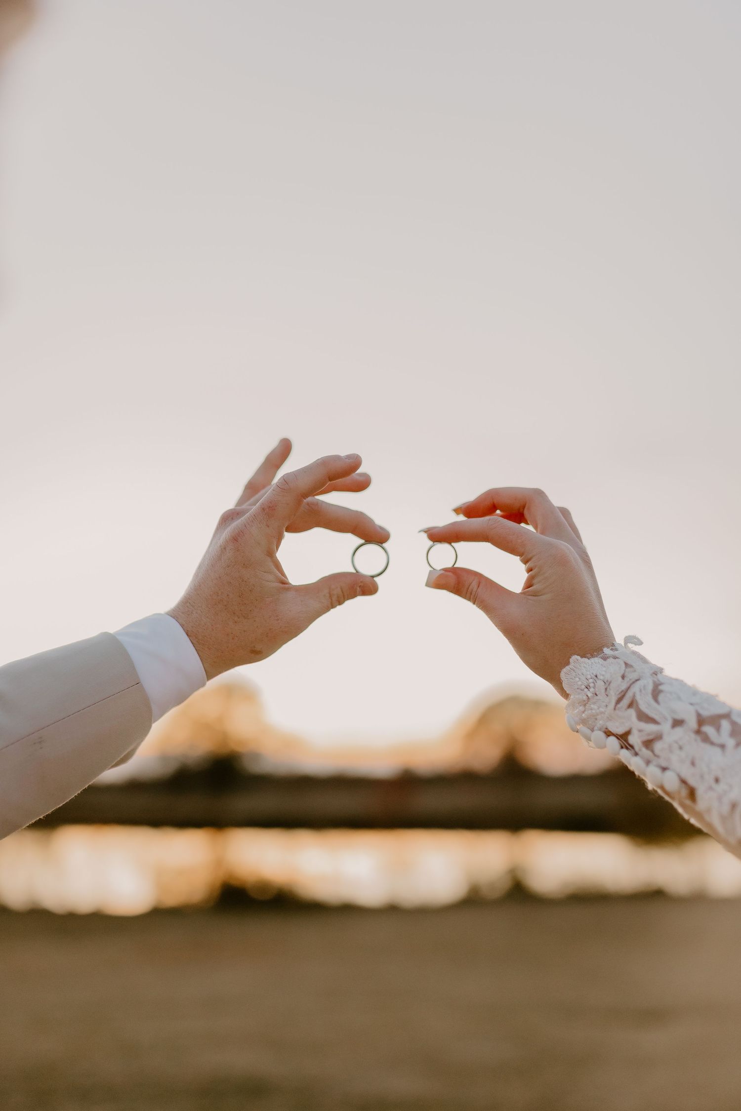Bride and groom holding wedding rings together at sunset, lace sleeve and suit visible in romantic outdoor ceremony.
