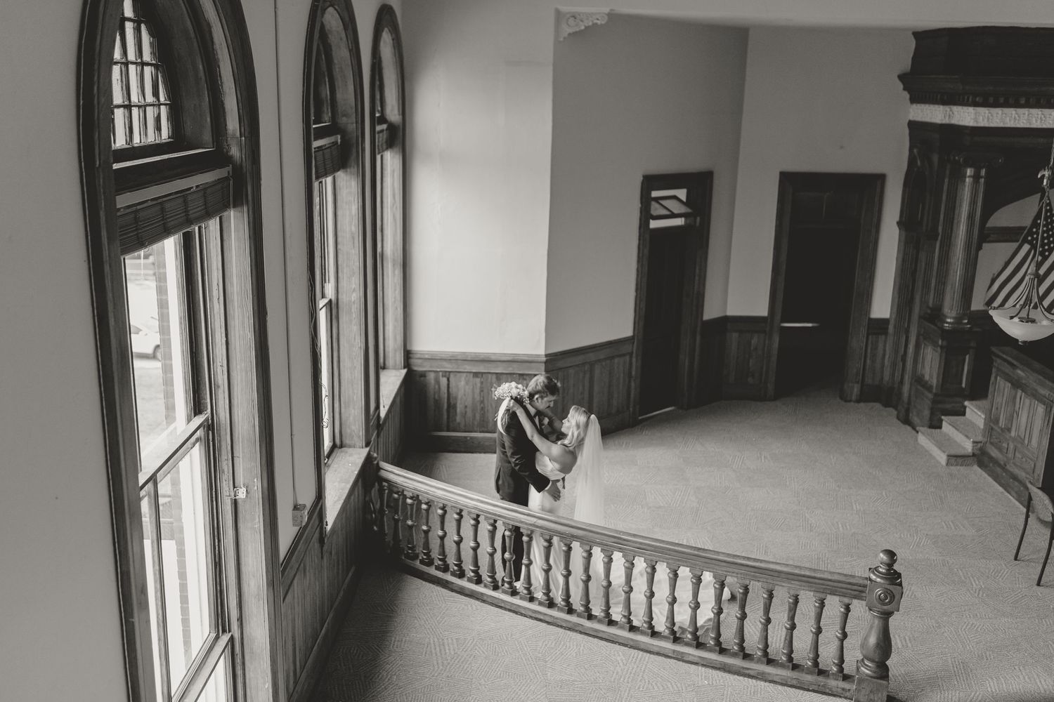 Black and white photo of a couple sharing a romantic kiss on an elegant staircase in a historic venue.
