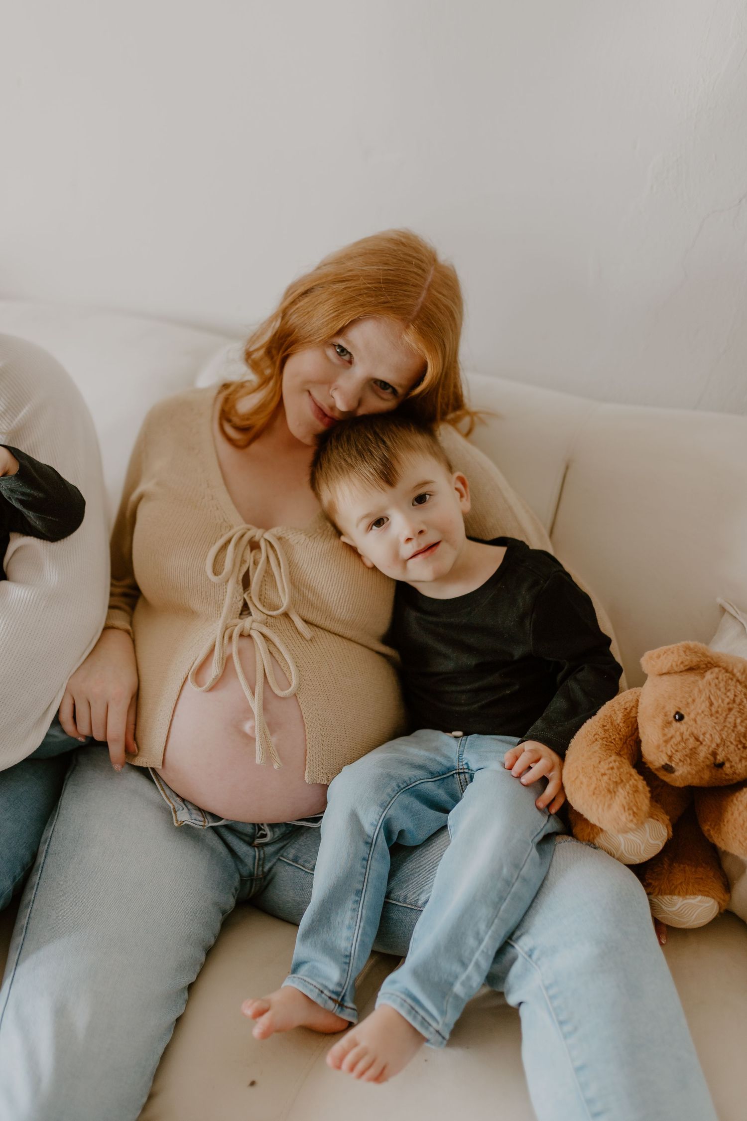 Pregnant woman with red hair sits on white sofa with young boy and teddy bear in cozy home setting.
