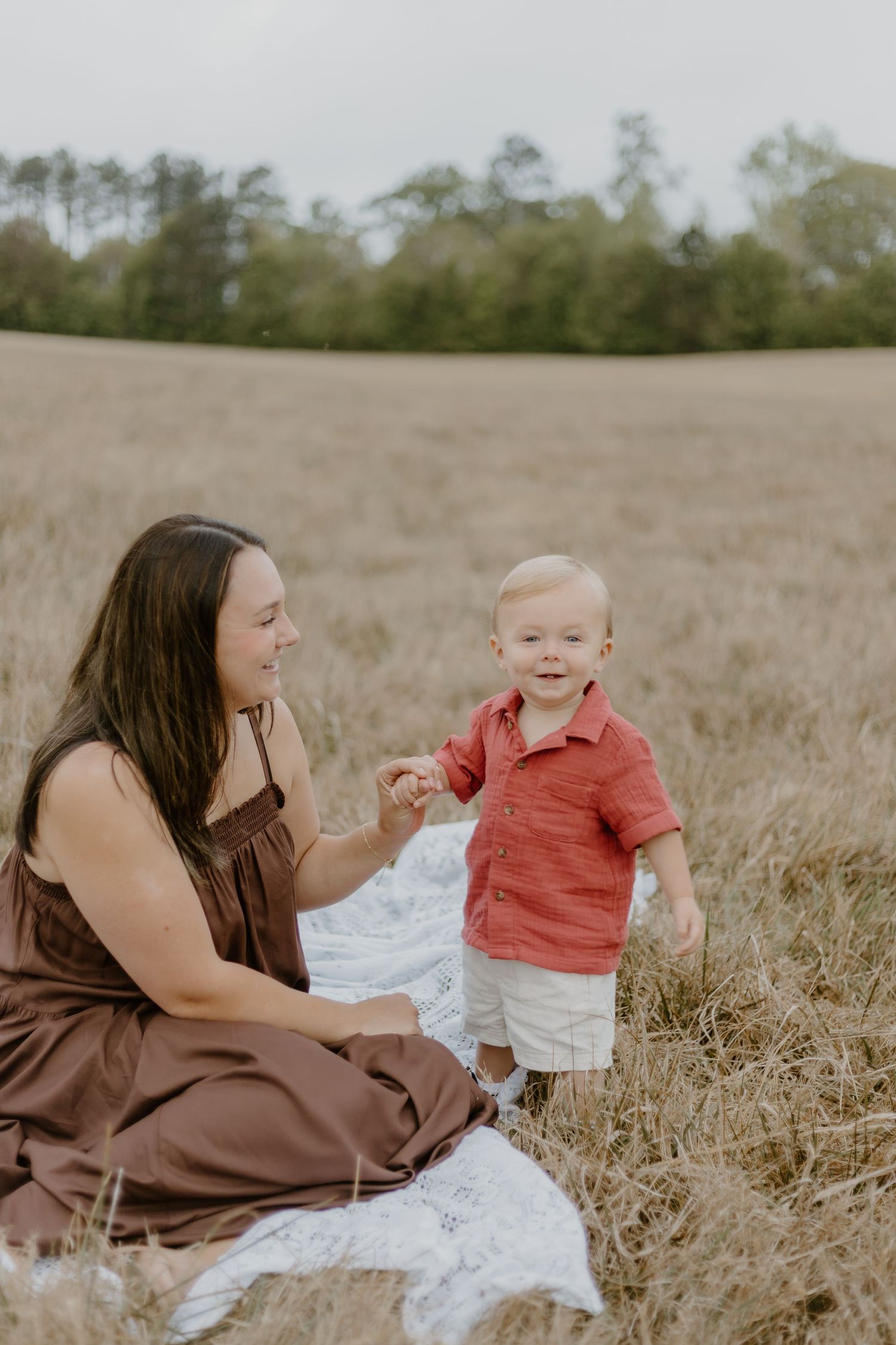 A mother in a brown dress sits on a blanket in a field, smiling at her toddler boy in a red shirt.