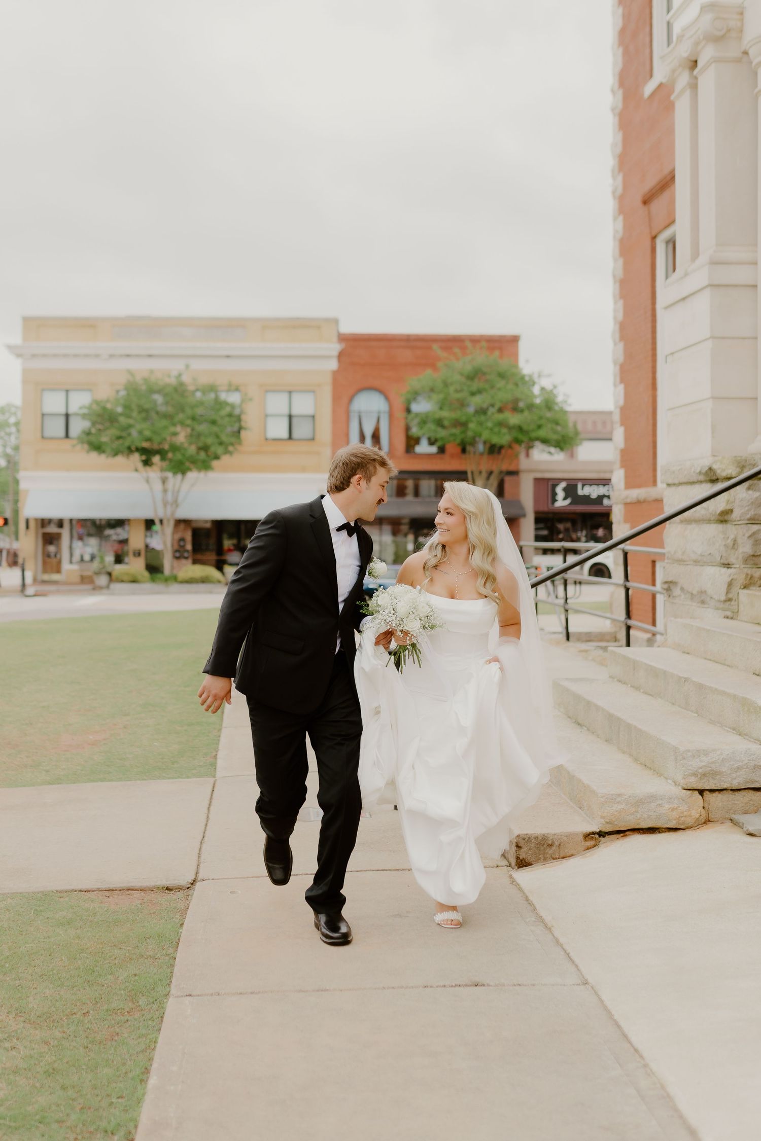 Bride and groom walking together outside a brick building, bride holding a bouquet in a white gown.
