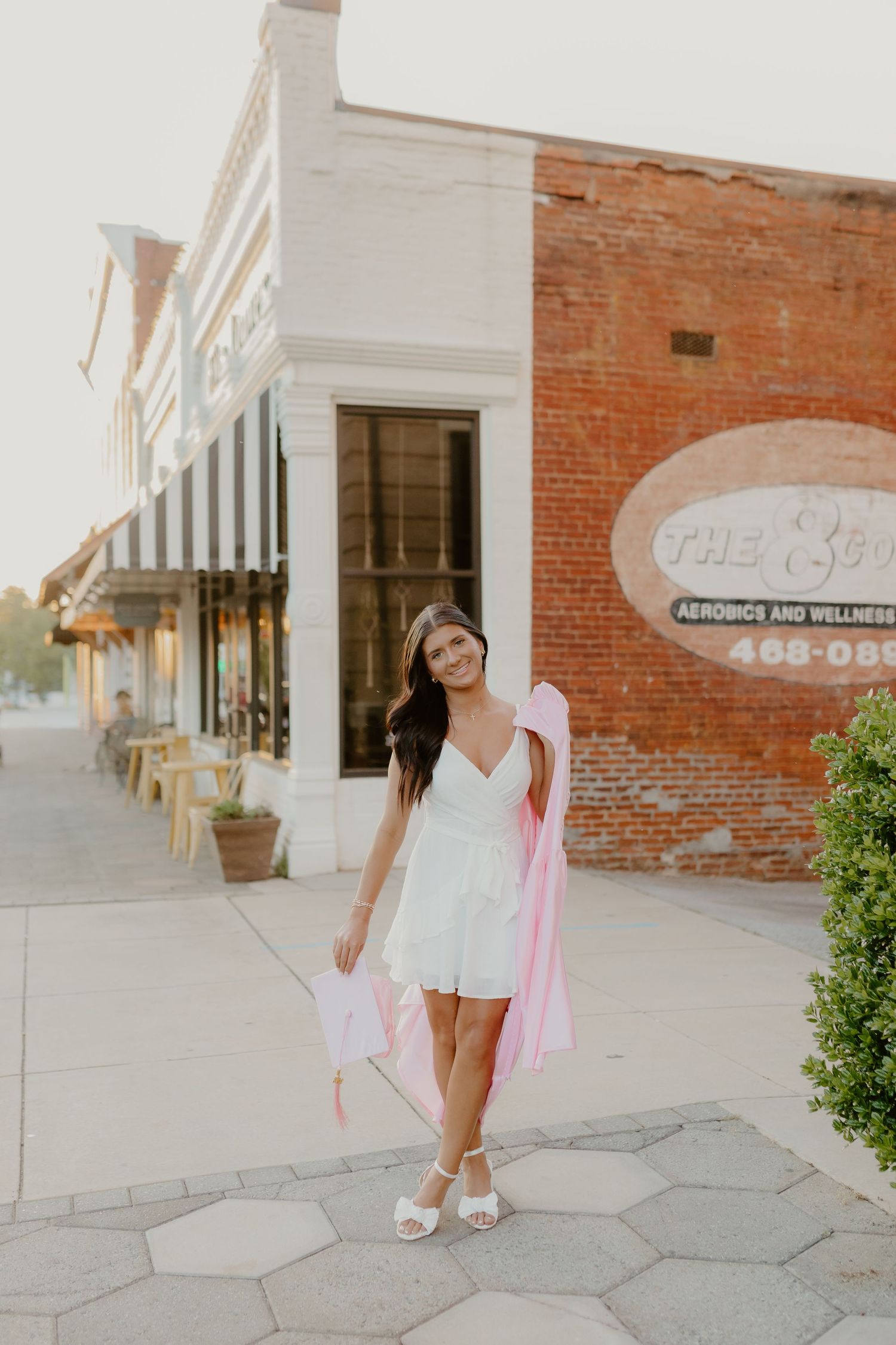 Young woman in white romper holding pink ribbon walks on brick sidewalk outside aerobics wellness center.