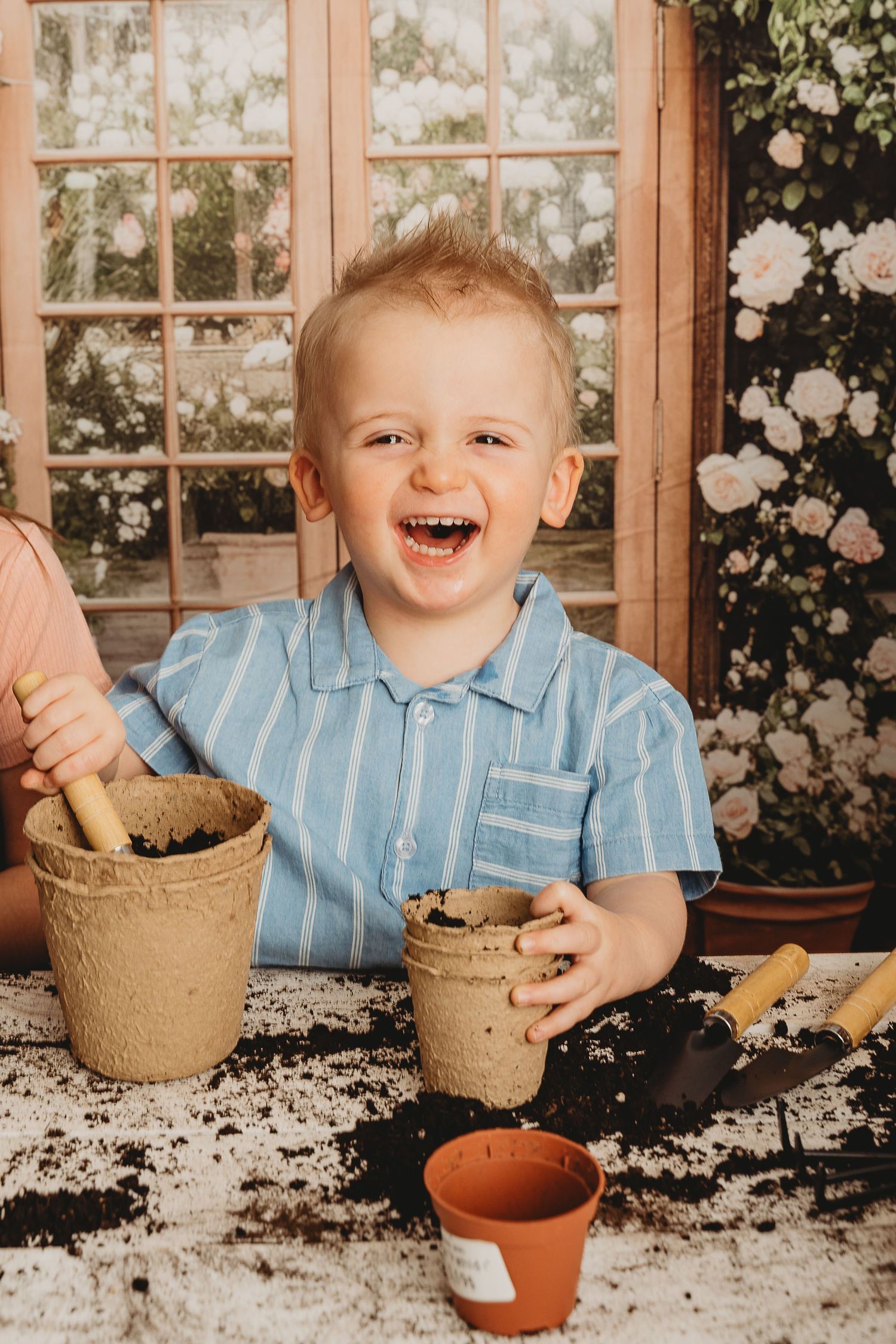 A laughing toddler boy in a blue striped shirt plays with soil and peat pots at a table with garden tools.
