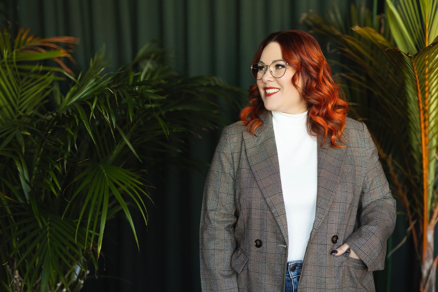 A smiling person with red curly hair wearing a gray plaid blazer and white turtleneck poses against a backdrop of tropical plants.