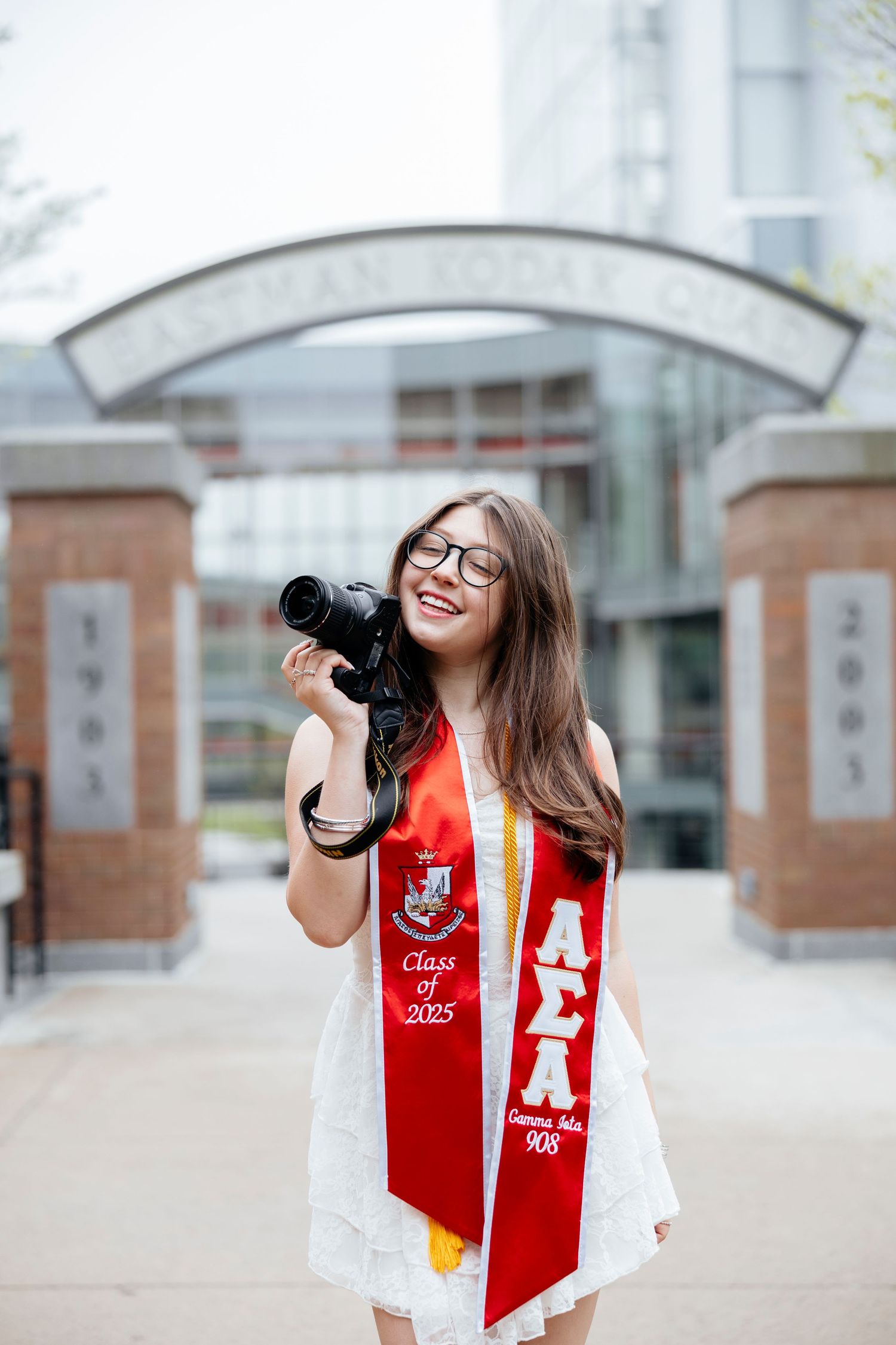Graduate in white dress and red stole taking photos with a professional camera outdoors during grad photos.