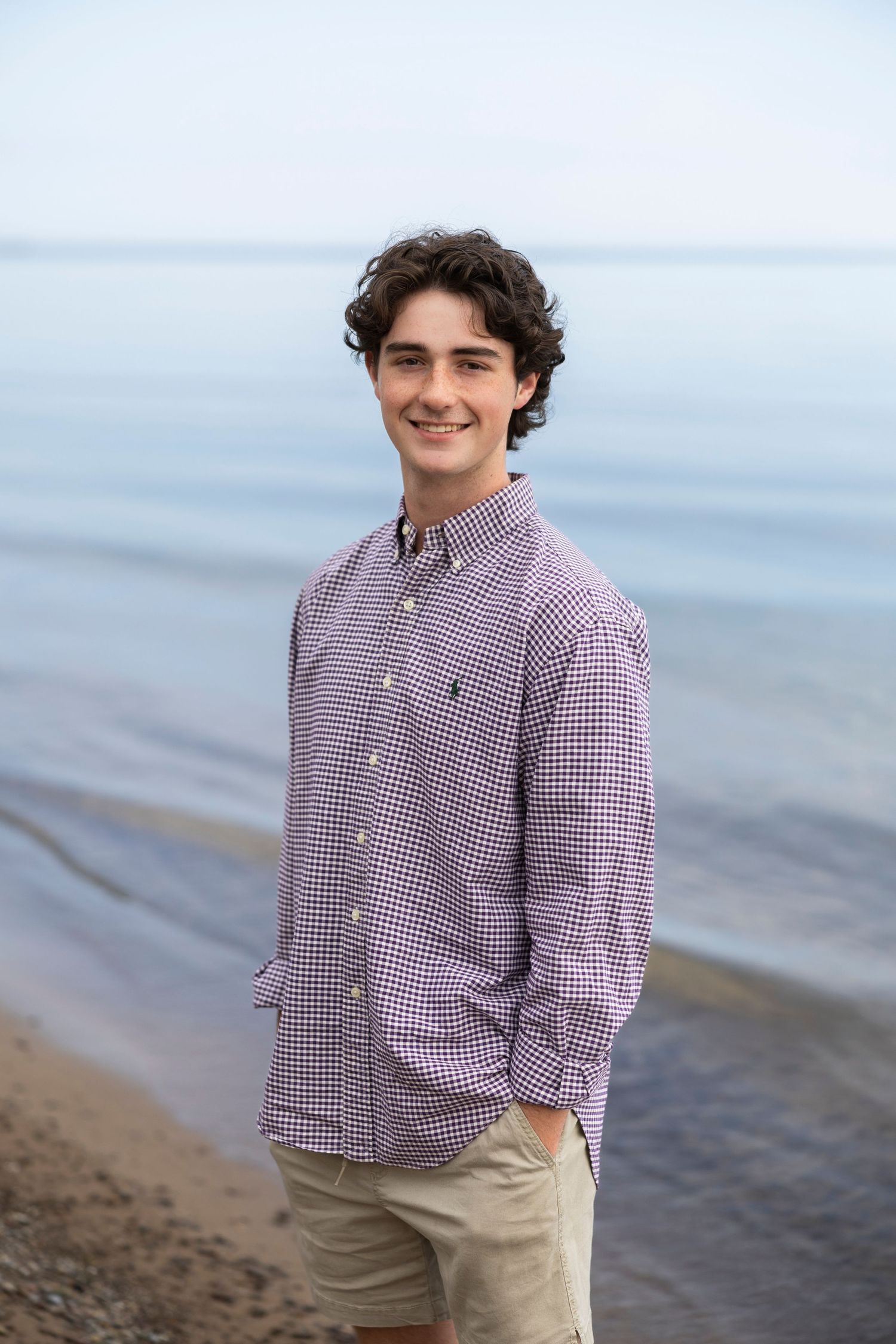 High school senior boy in red and white checkered shirt poses casually on Durand beach with ocean backdrop, Rochester, NY.