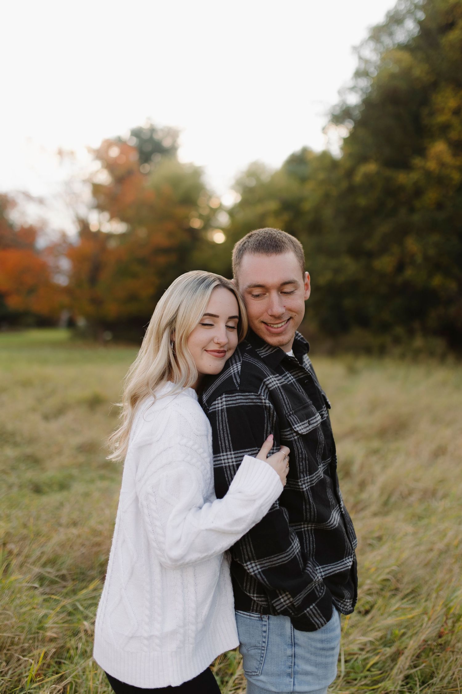 A couple sharing a joyful moment in a fall field with autumn foliage in the background at Rochester, NY couples photoshoot.