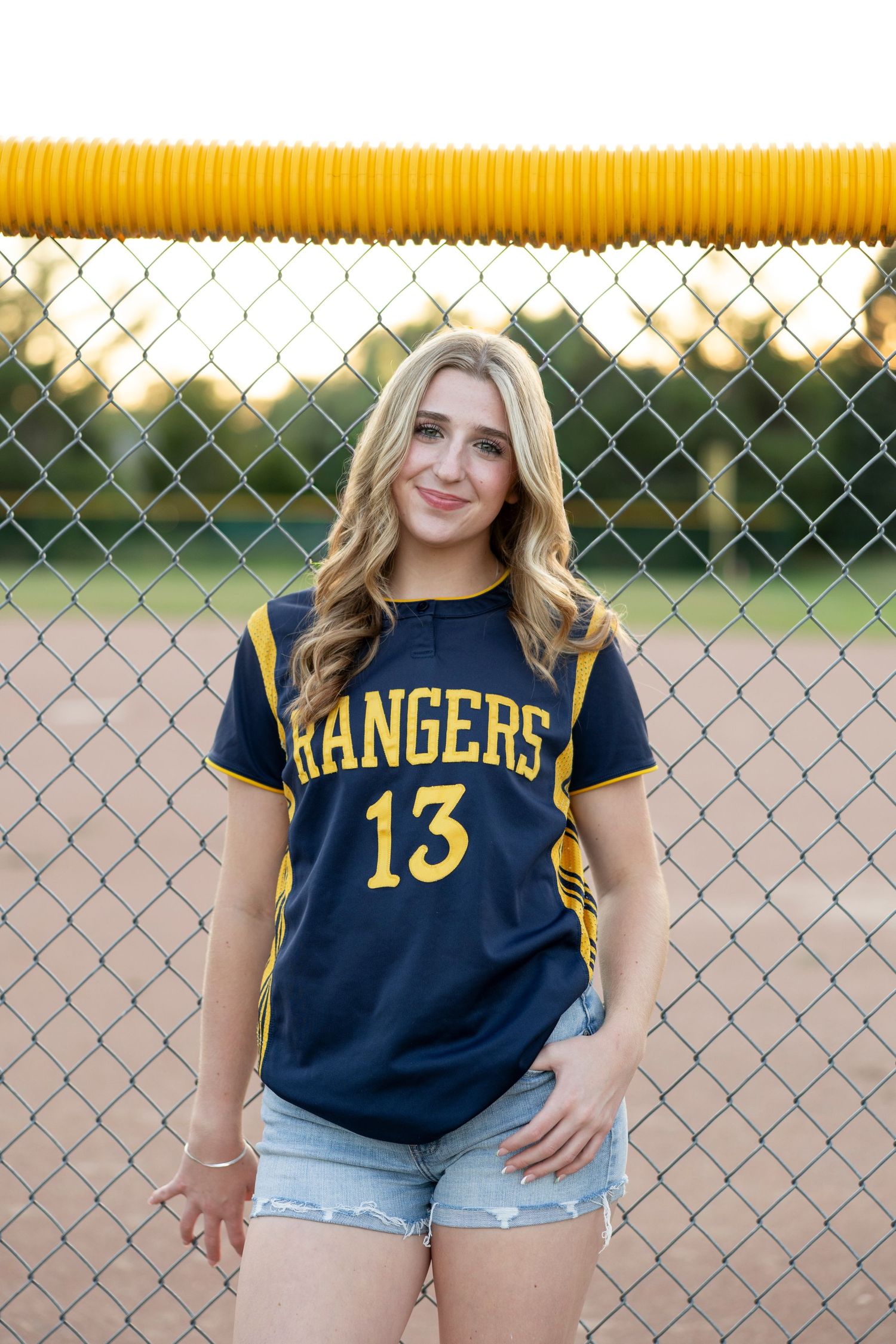 Softball sports themed portrait with Spencerport Rangers jersey and denim shorts by chain link fence. Rochester, NY senior portaits.