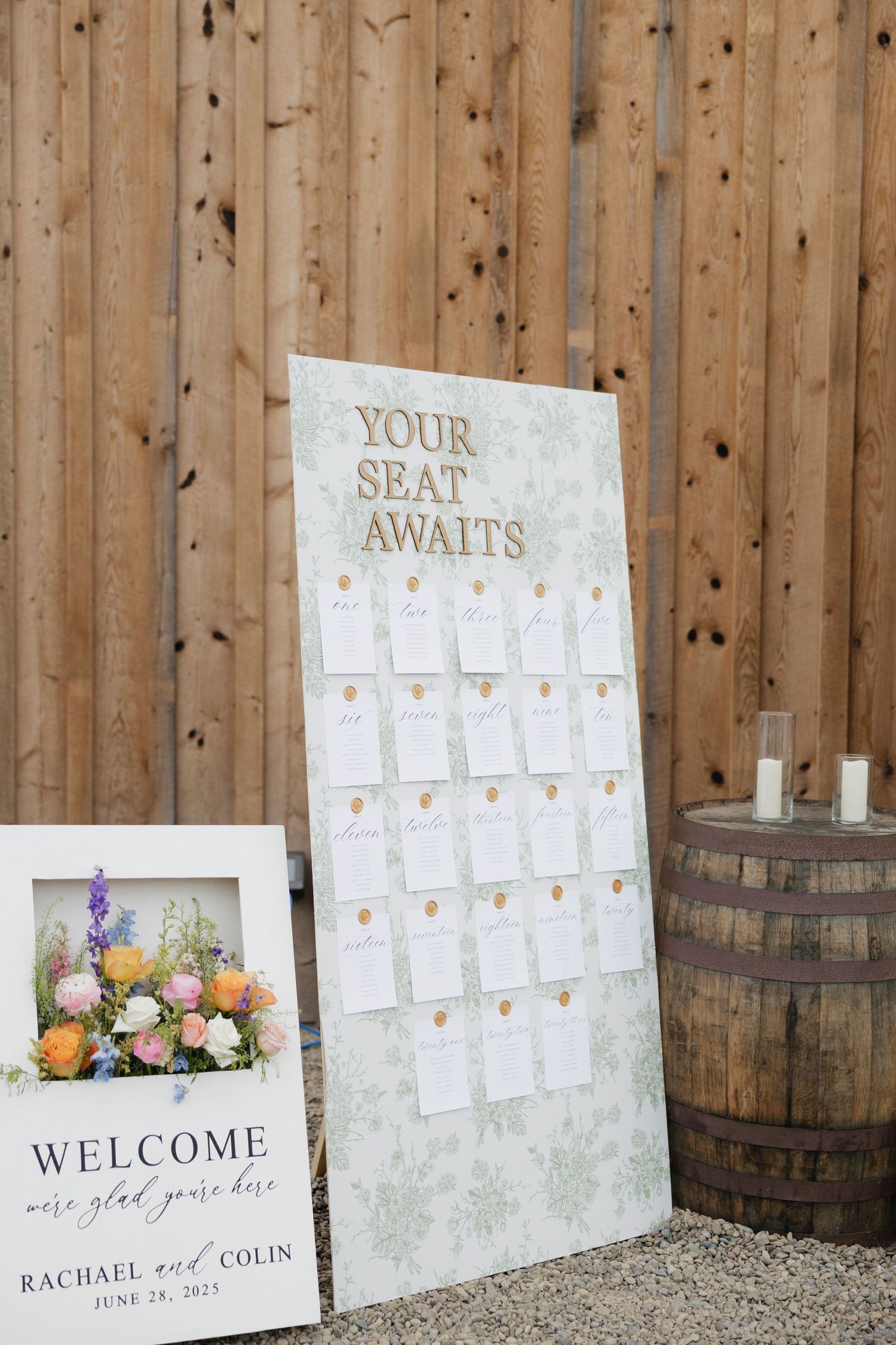 Rustic wedding seating chart display against wooden barn with wine barrels and floral arrangements in Rochester, NY barn wedding.