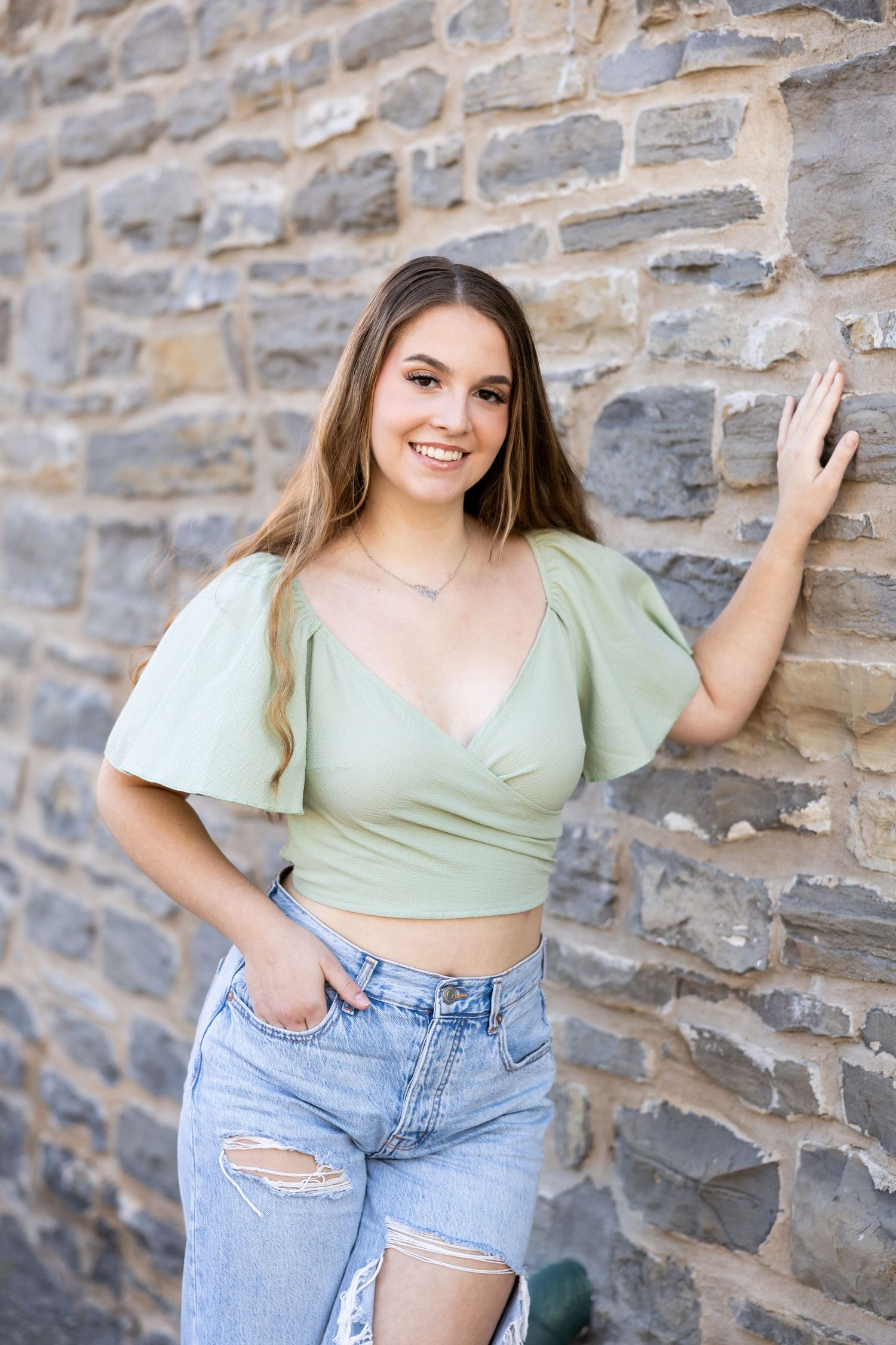 Blonde high school senior girl in mint green crop top and denim shorts poses against stone wall for senior photo pose in Rochester, NY.
