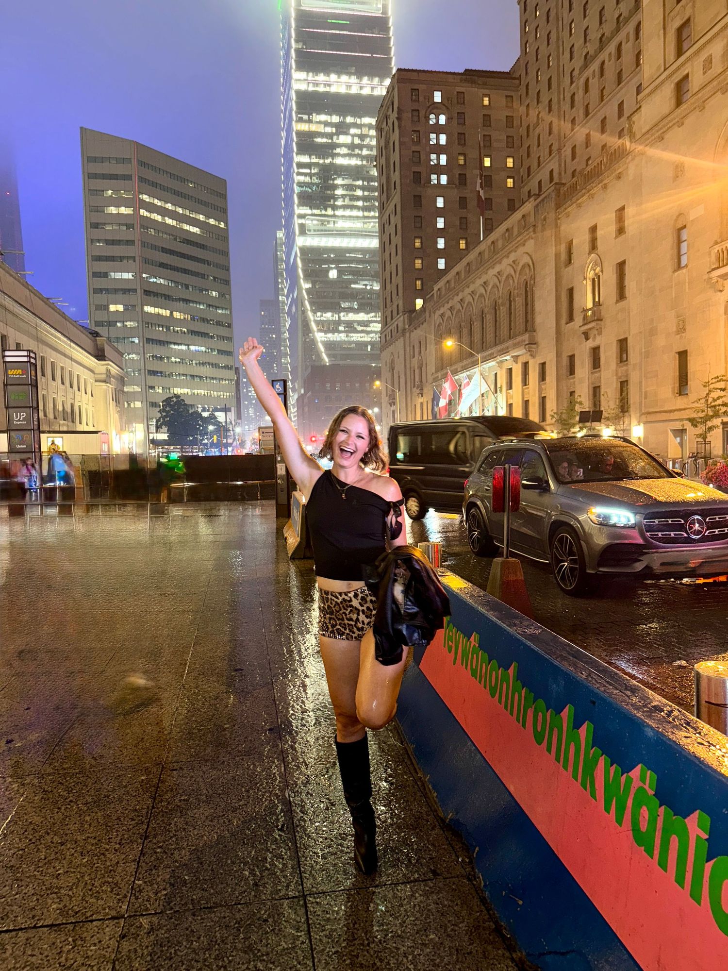 Woman in black top and leopard shorts poses joyfully in rainy city street at night with skyscrapers.