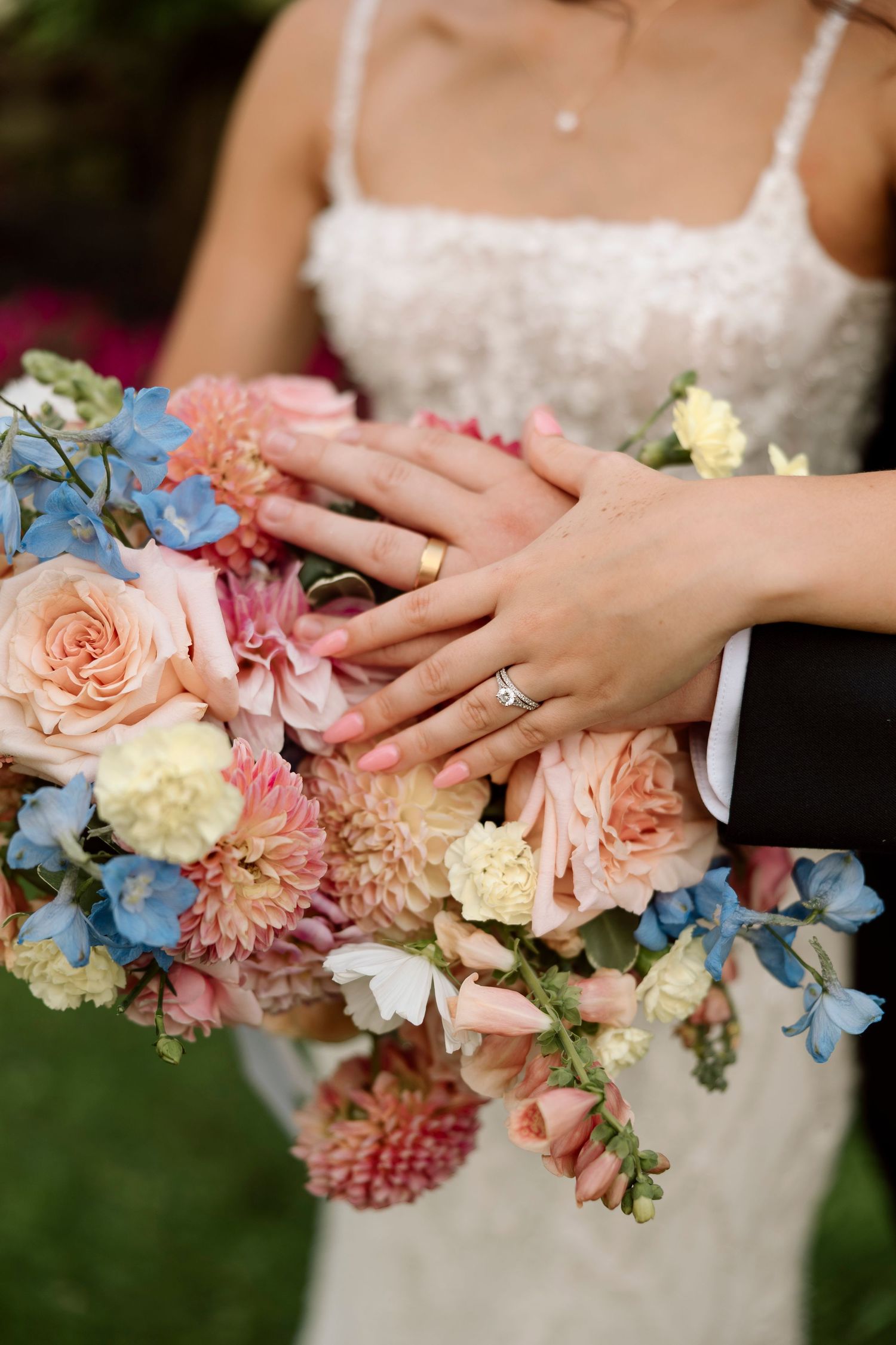 Close-up of hands with engagement ring resting on pastel bouquet with roses and blue delphiniums at Shadow Lake wedding Rochester, NY.