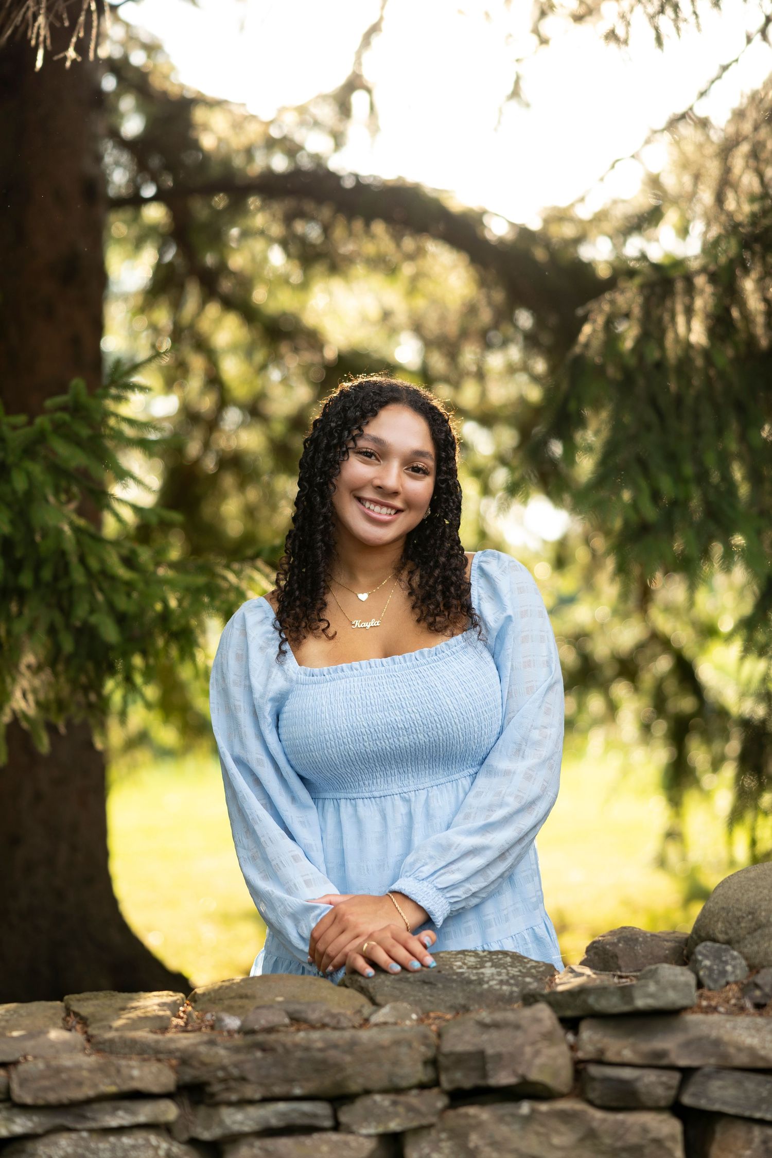 High school senior girl in light blue peasant top smiles while leaning on stone wall with trees in background at Saras Garden, Brockport.