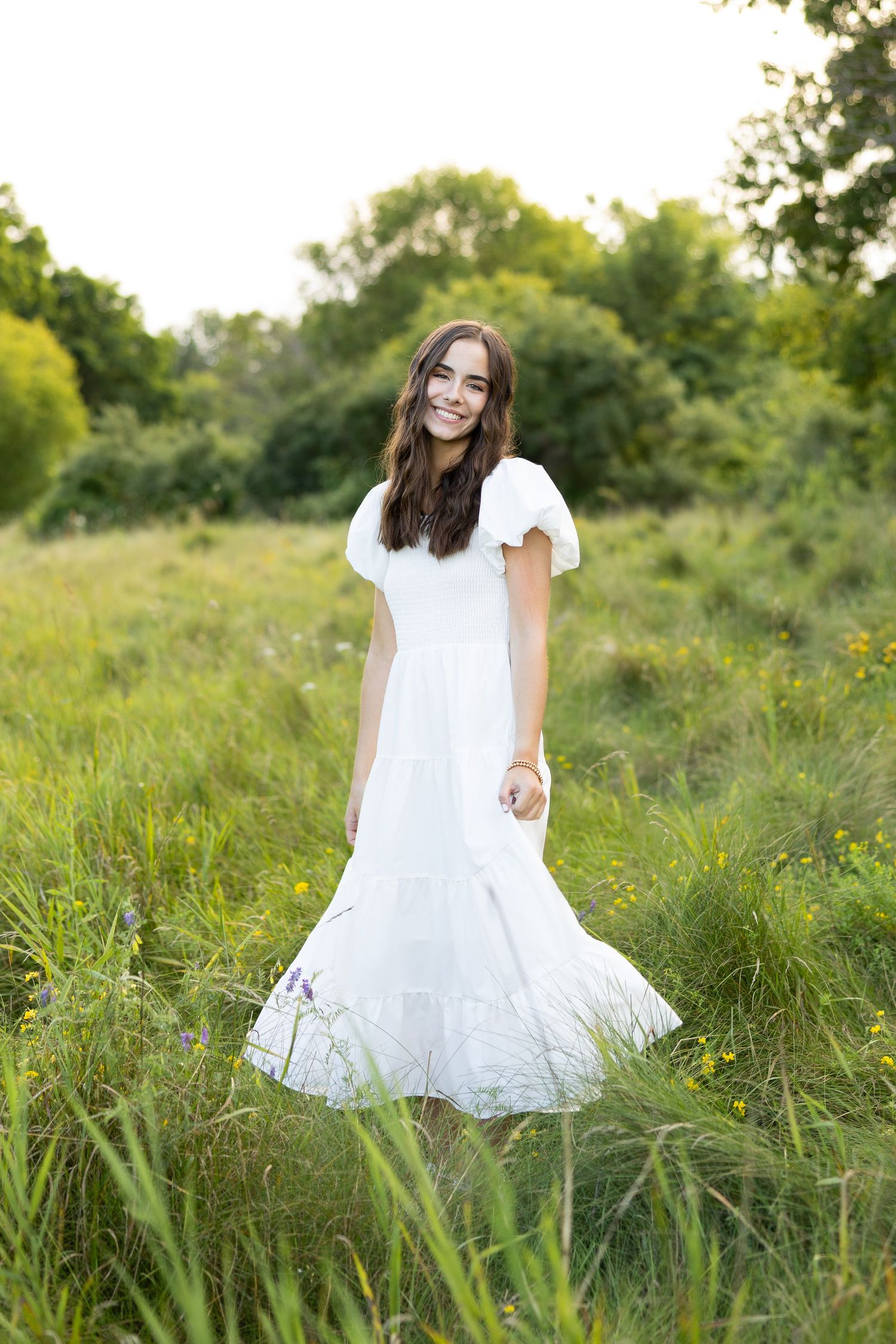 High school senior girl in flowing white dress twirls in tall grass field during golden hour at Hamlin Beach, Hamlin NY.