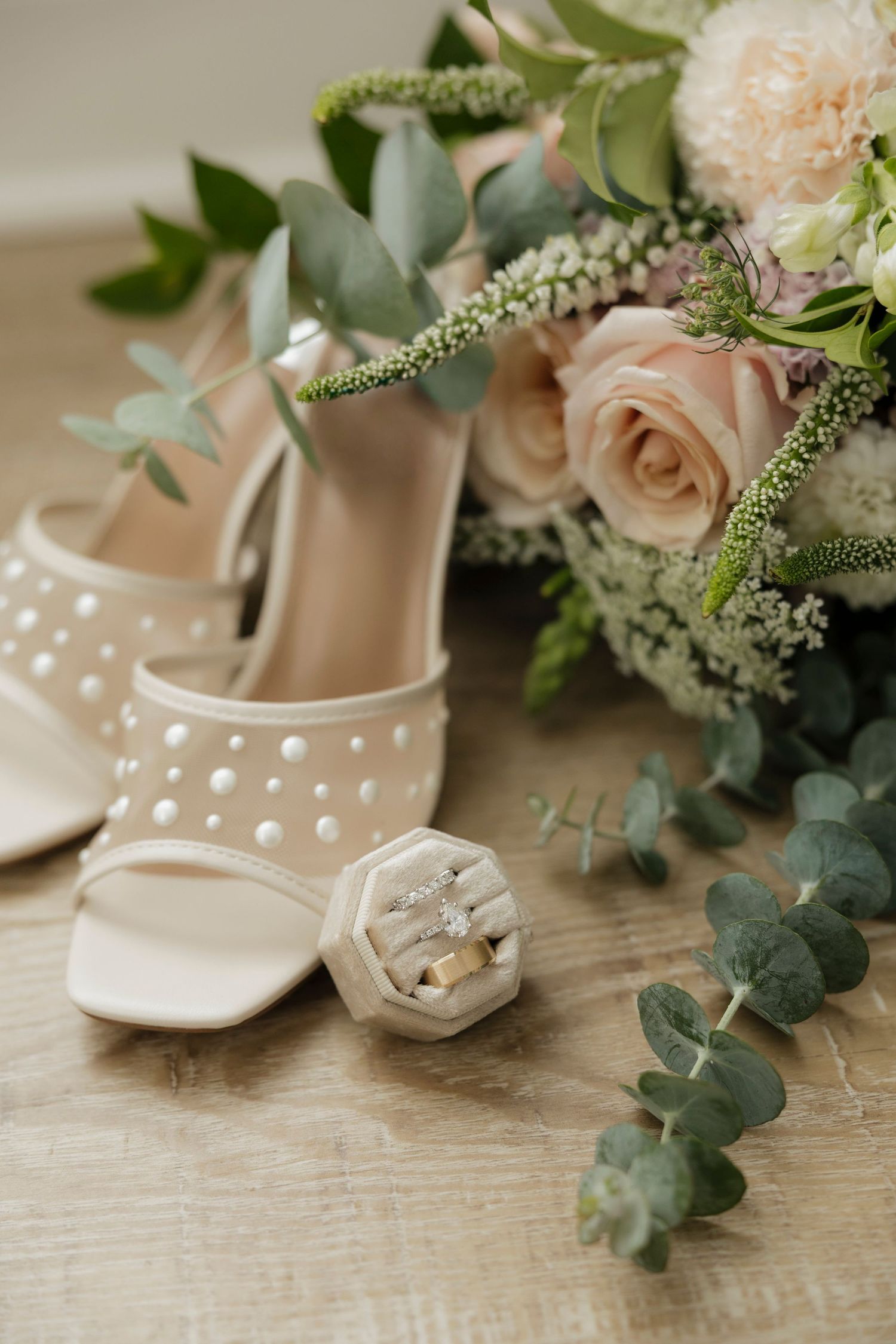 Soft pink pastel wedding accessories including beige heels, rings, and delicate floral arrangements with eucalyptus.